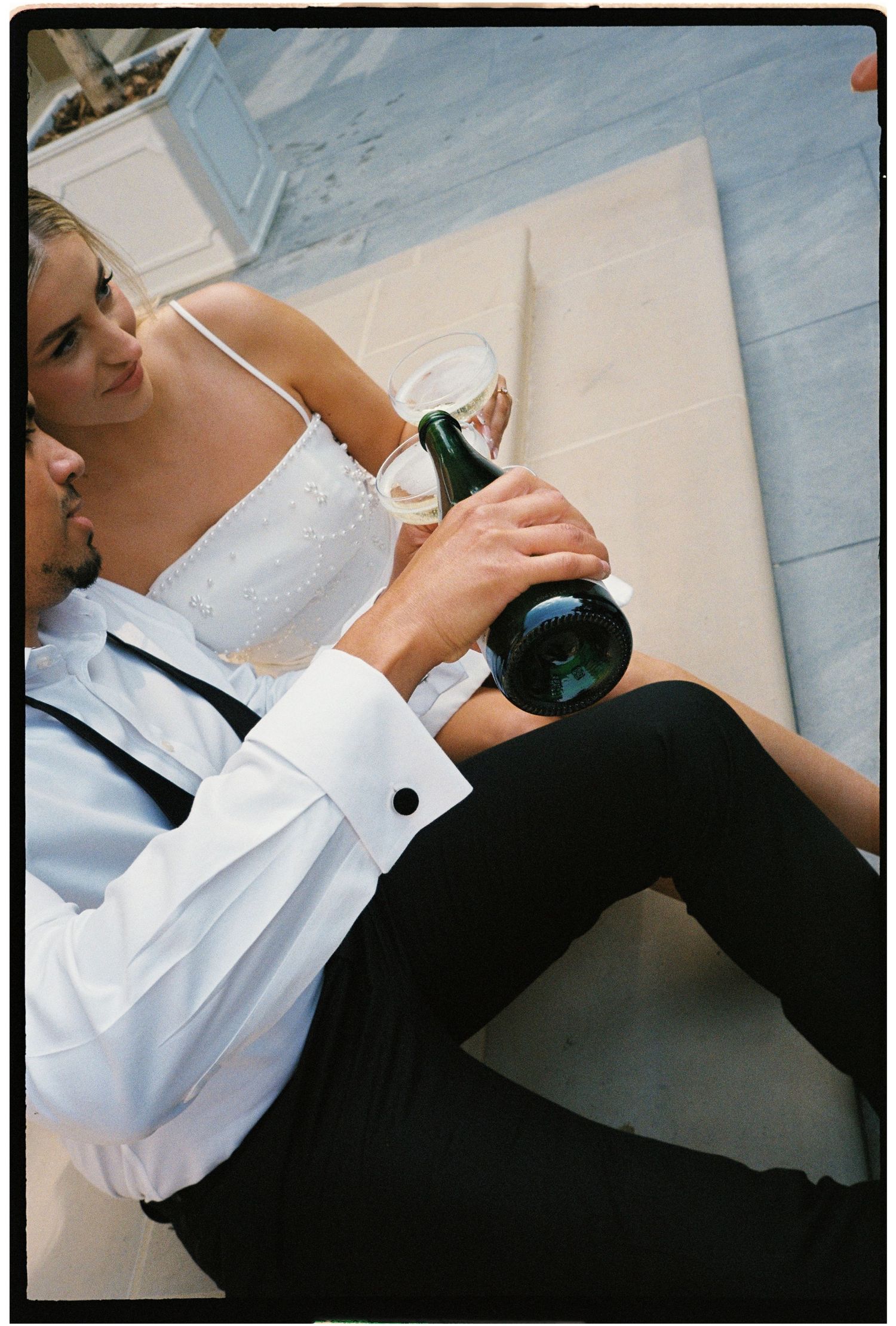 Couple in formal wear sitting on floor, man pouring champagne into glass, wedding celebration moment.