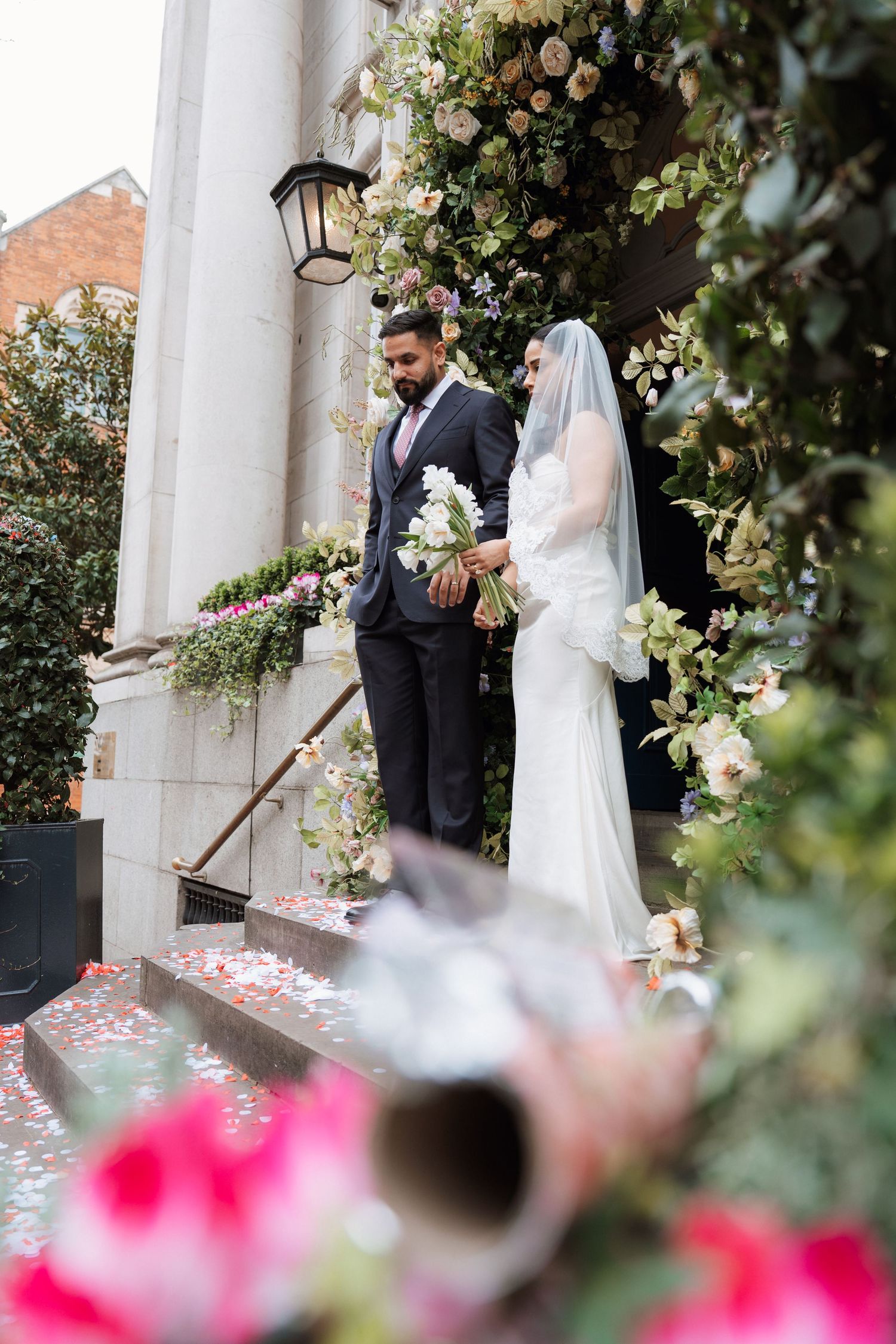 A bride and groom share a romantic moment on steps adorned with pink rose petals and floral decorations.