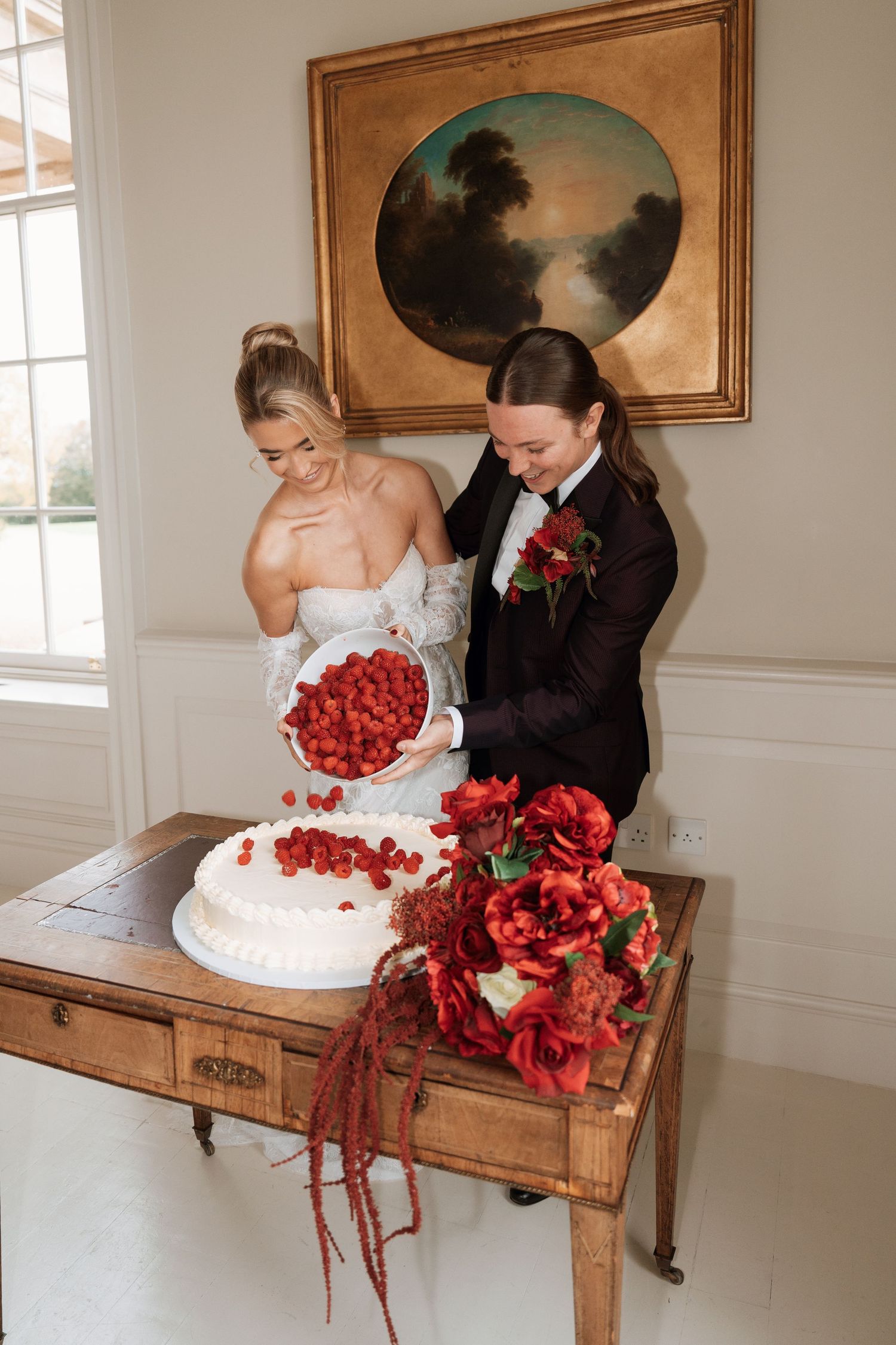 Wedding couple cutting cake decorated with red flowers on antique wooden table.