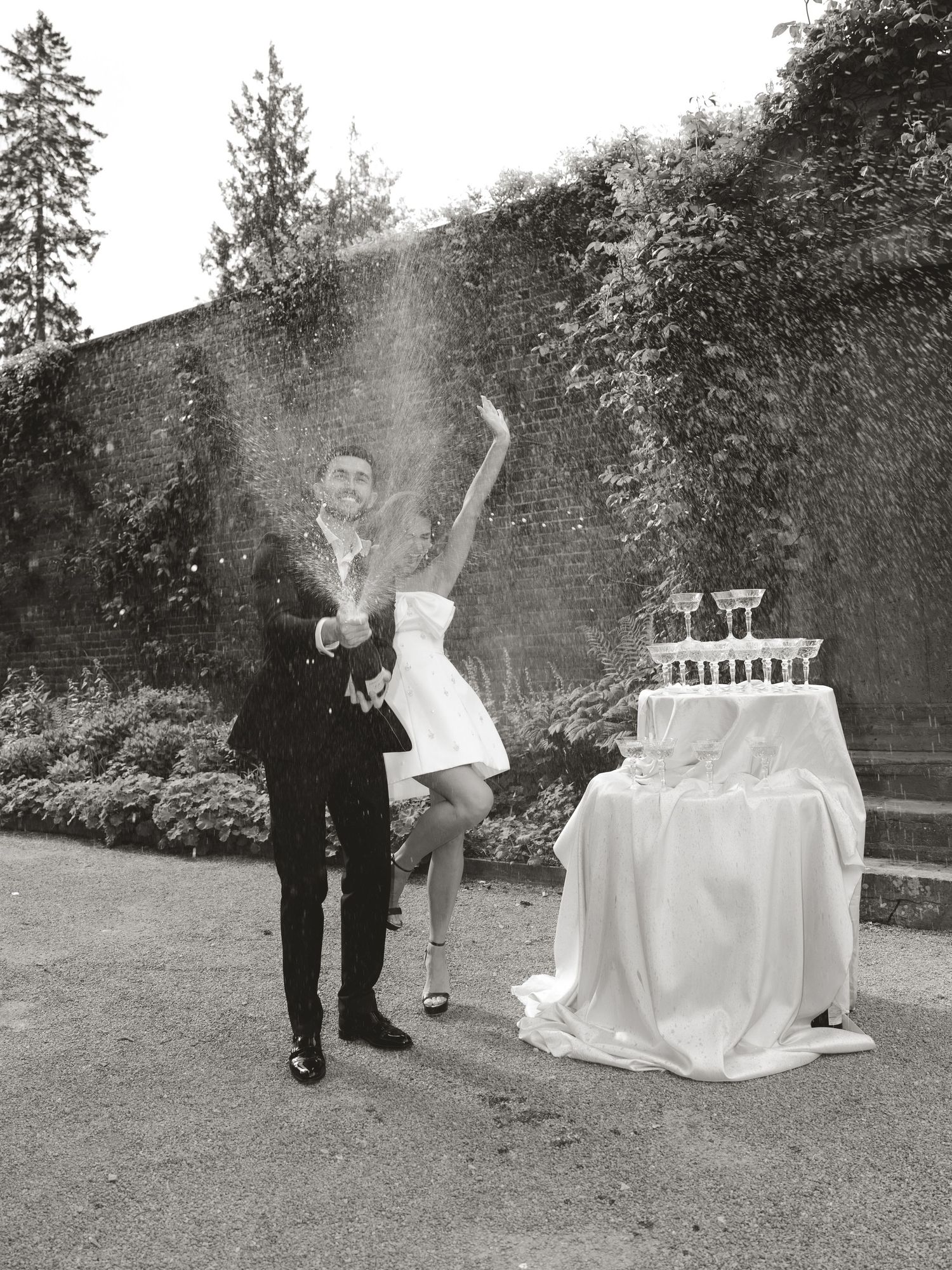 Black and white photo of a bride and groom celebrating with champagne tower at outdoor wedding reception.