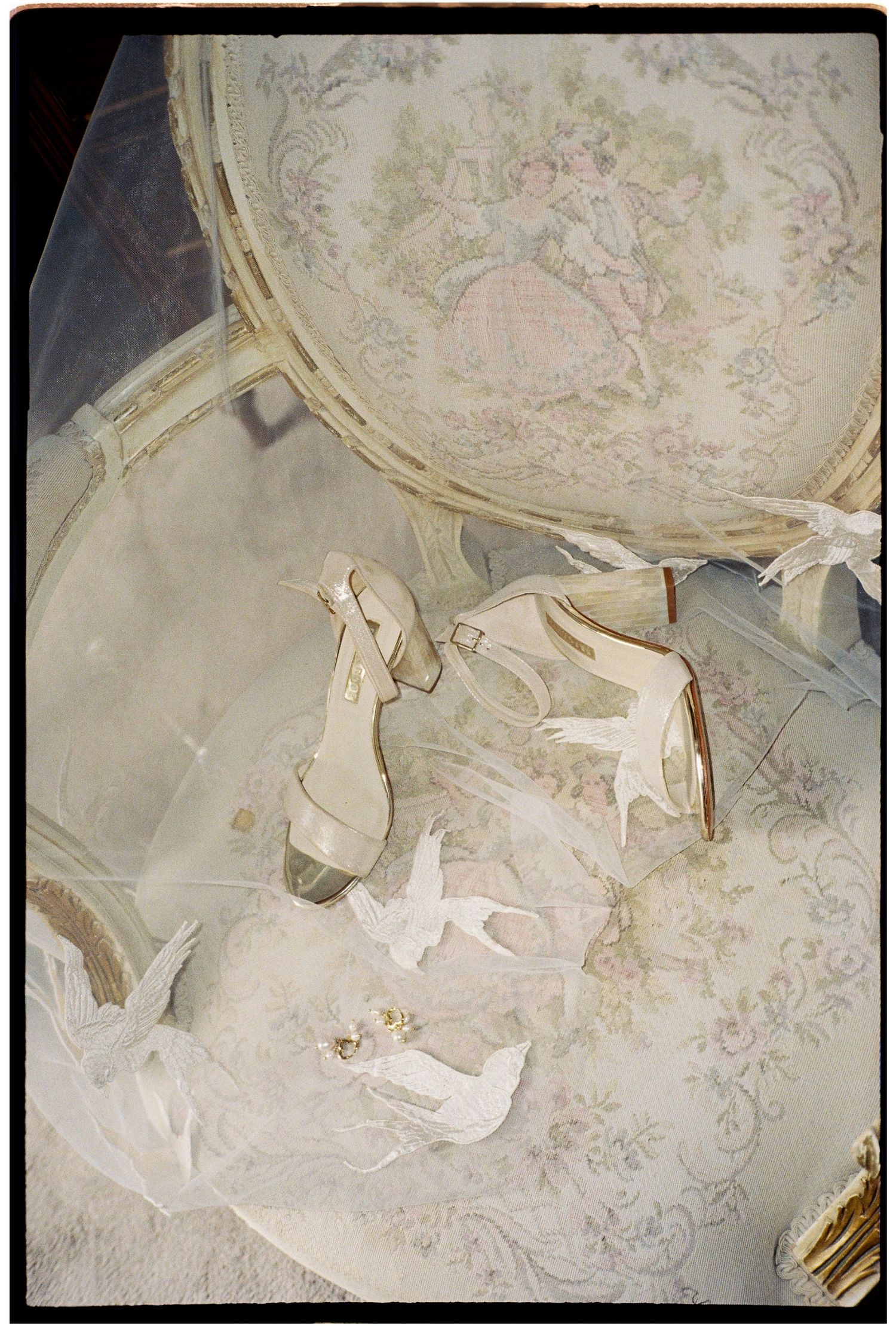 Elegant bridal heels with ribbon ties rest on a floral tapestry chair alongside a delicate veil.