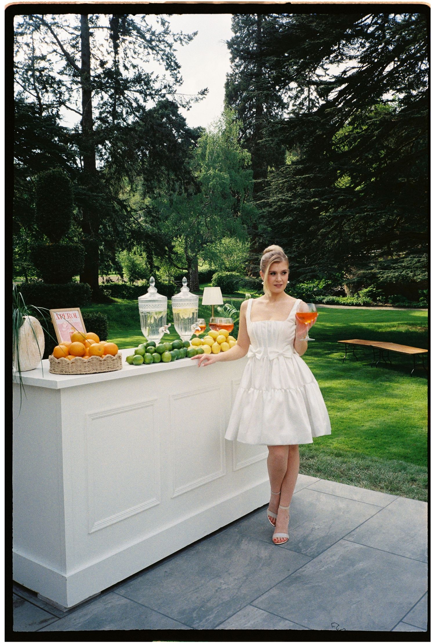 Woman in white dress holding citrus fruit stands beside outdoor bar with fresh fruits and drink dispensers in lush garden.