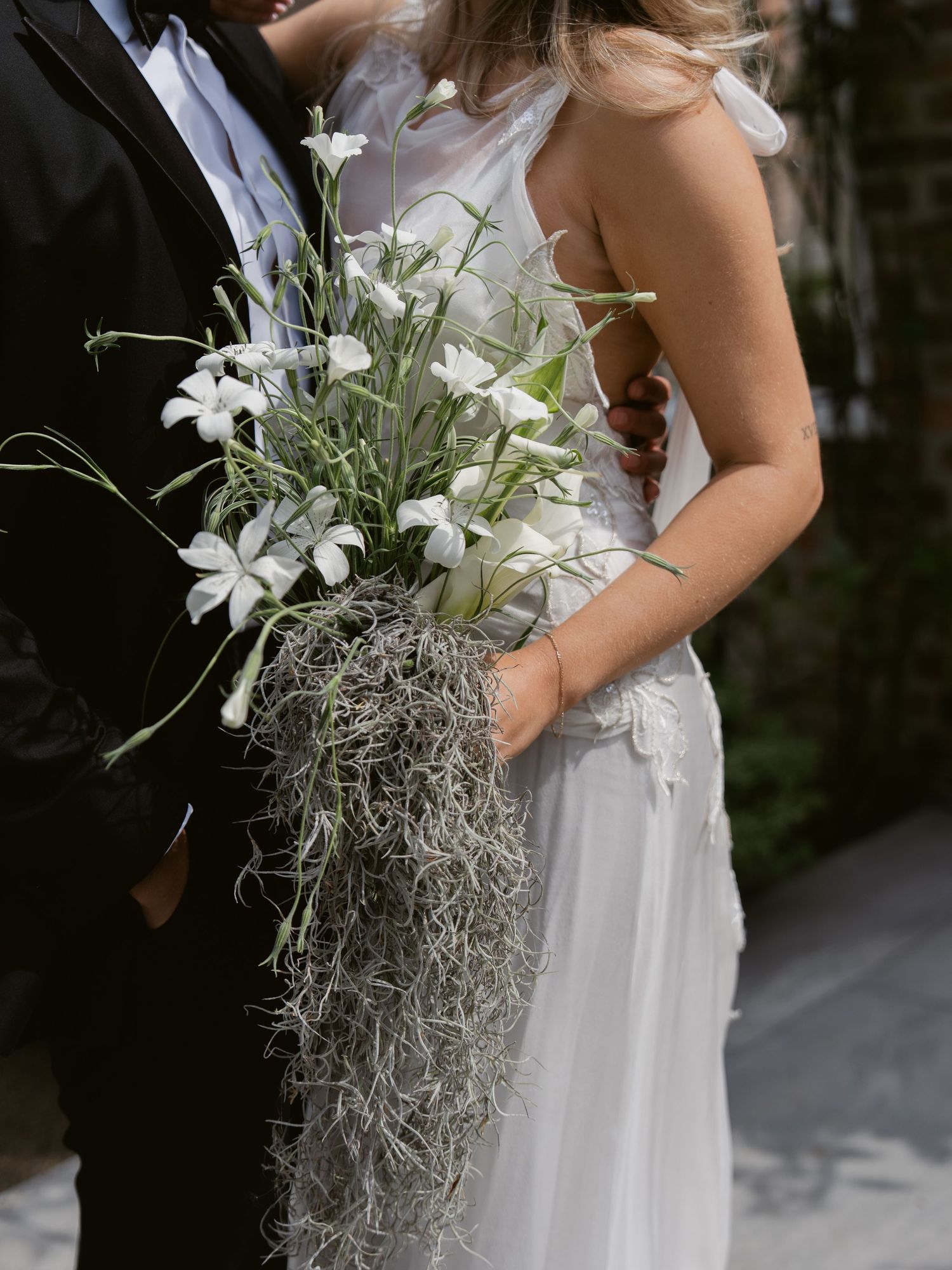 Bride holding a cascading white floral bouquet with Spanish moss, standing beside groom in black tuxedo.