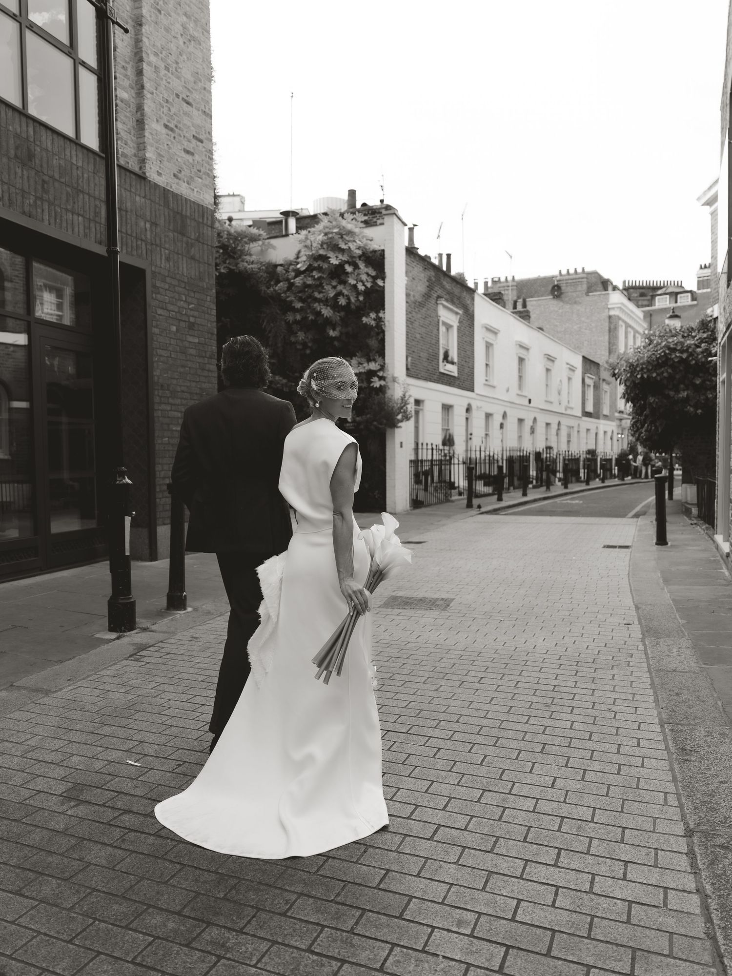 Black and white photo of a bride in a white gown and groom in a suit walking down a cobblestone street.