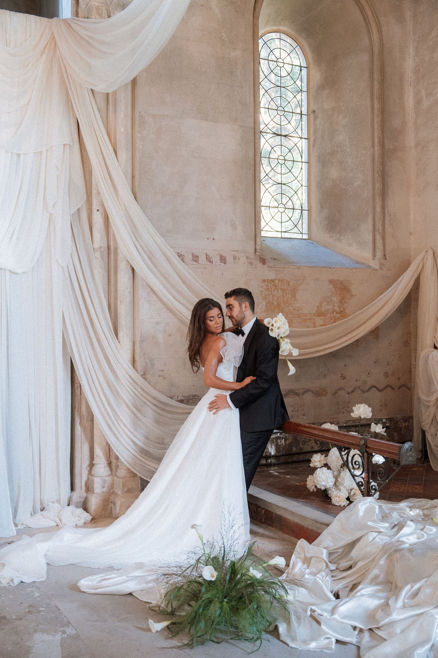 Wedding couple dancing in a historic chapel decorated with flowing white fabric and floral arrangements.