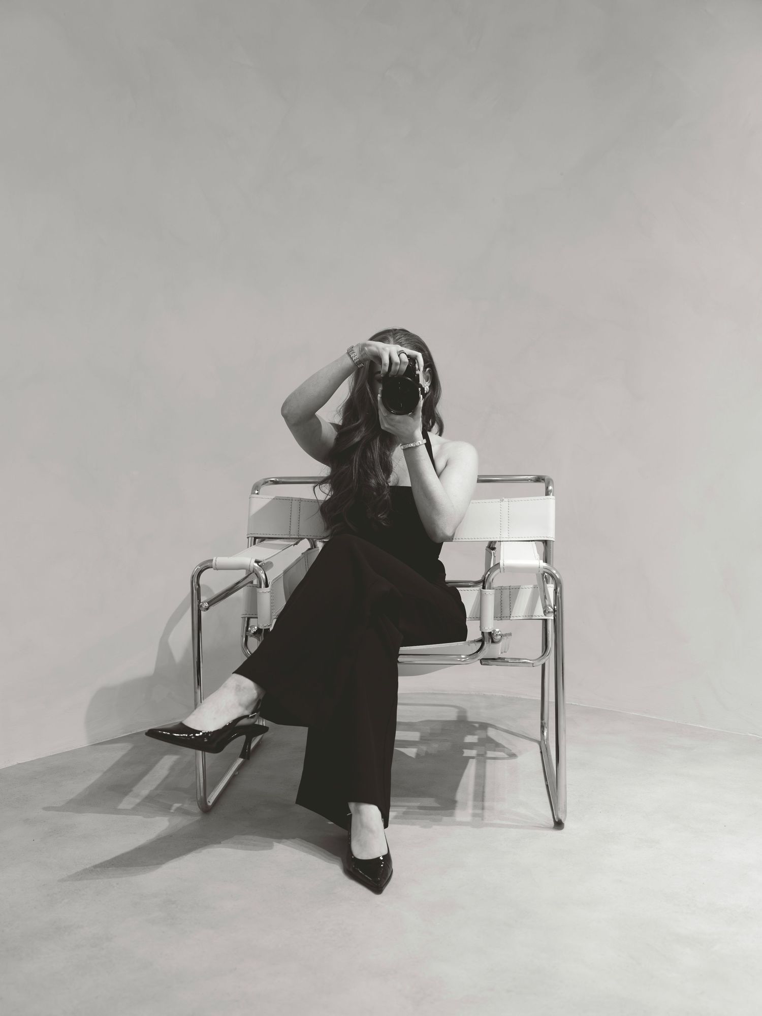 Woman in black outfit sits on modern chair, holding camera up to face, black and white studio photo.
