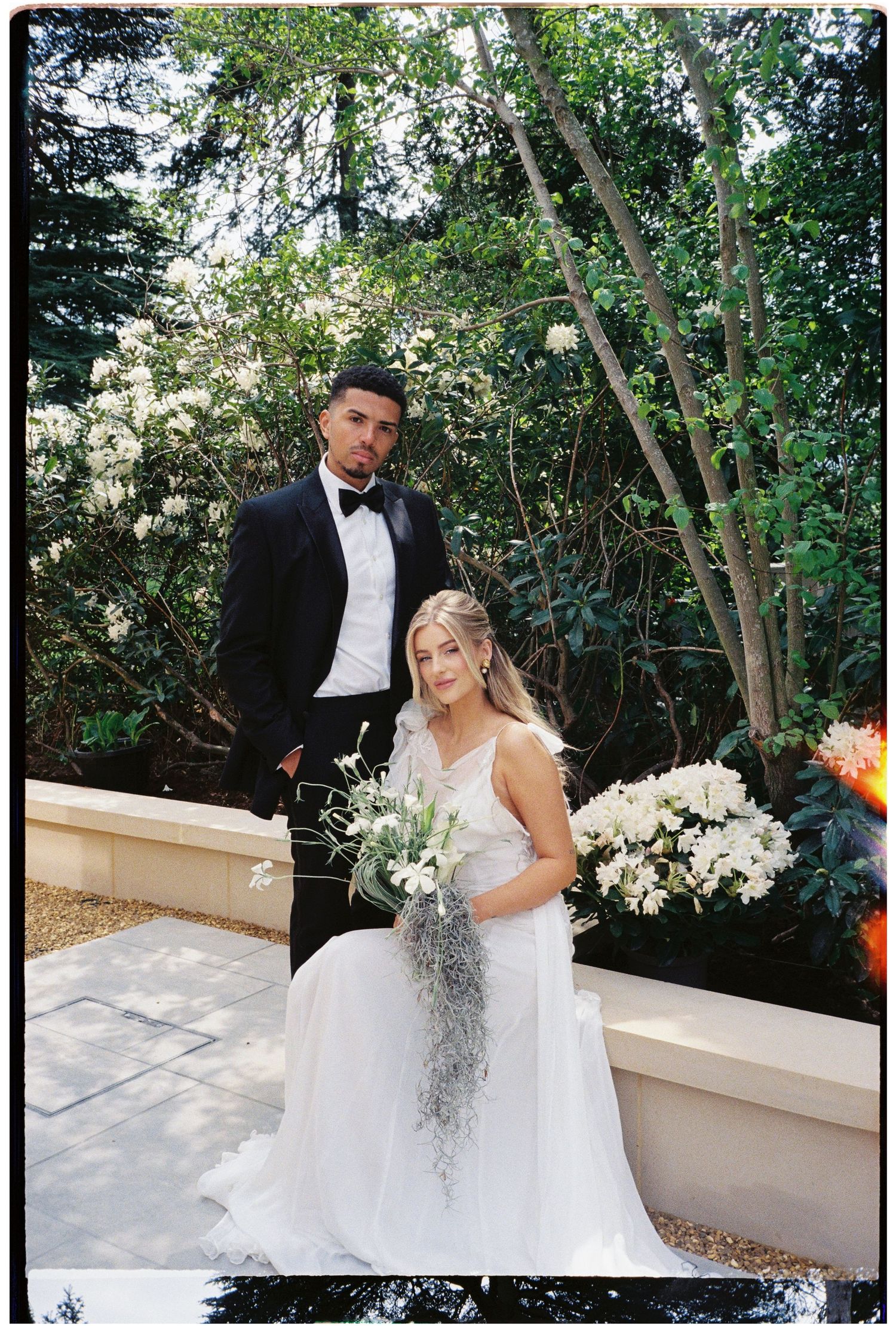Elegant wedding couple posing outdoors, groom in black tuxedo, bride in white gown holding cascading floral bouquet.