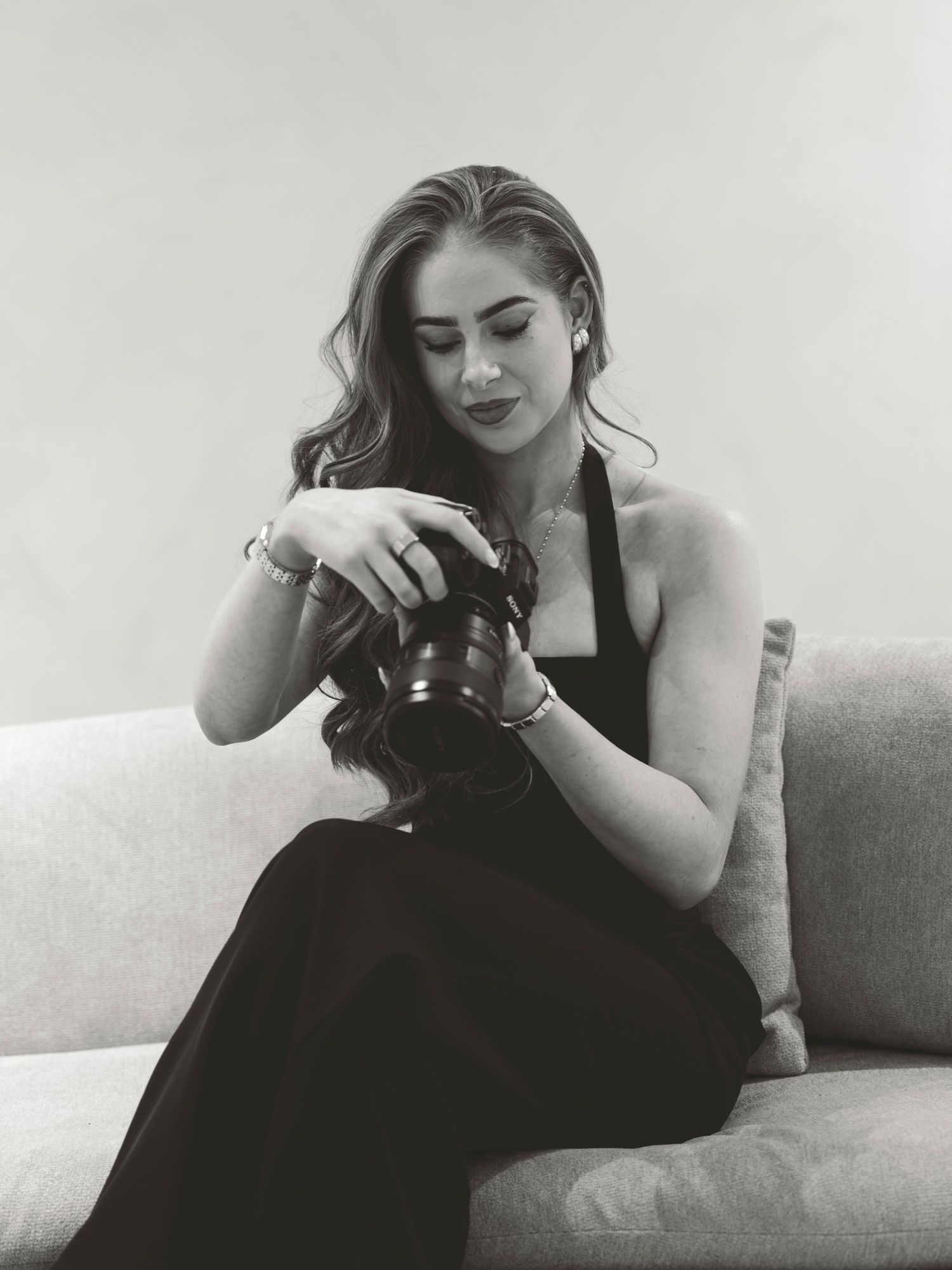 Black and white photo of a young woman in a halter dress sitting on a sofa, examining a camera.