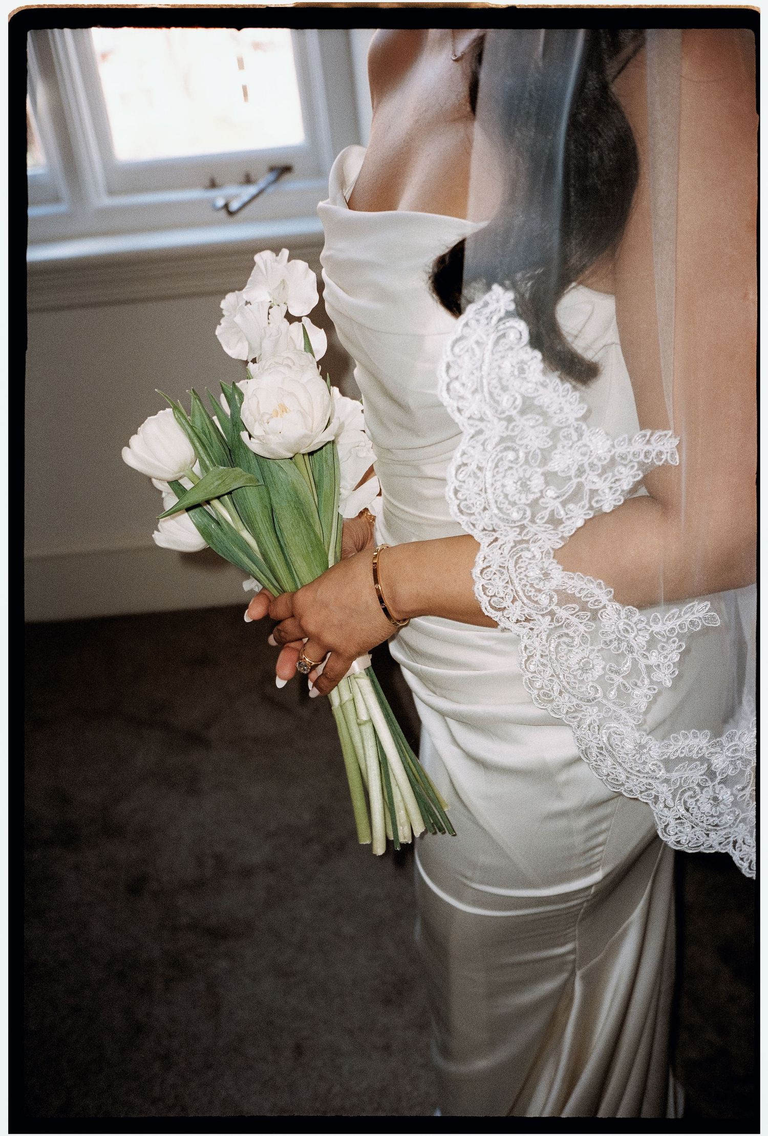 A bride in a white satin dress holds a simple bouquet of white tulips against a soft interior backdrop.
