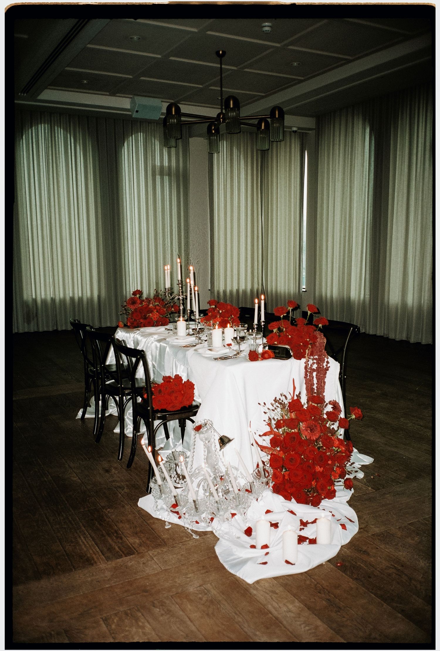 Elegant table setting with white linens and dramatic red floral arrangements spilling onto hardwood floor in dimly lit room.