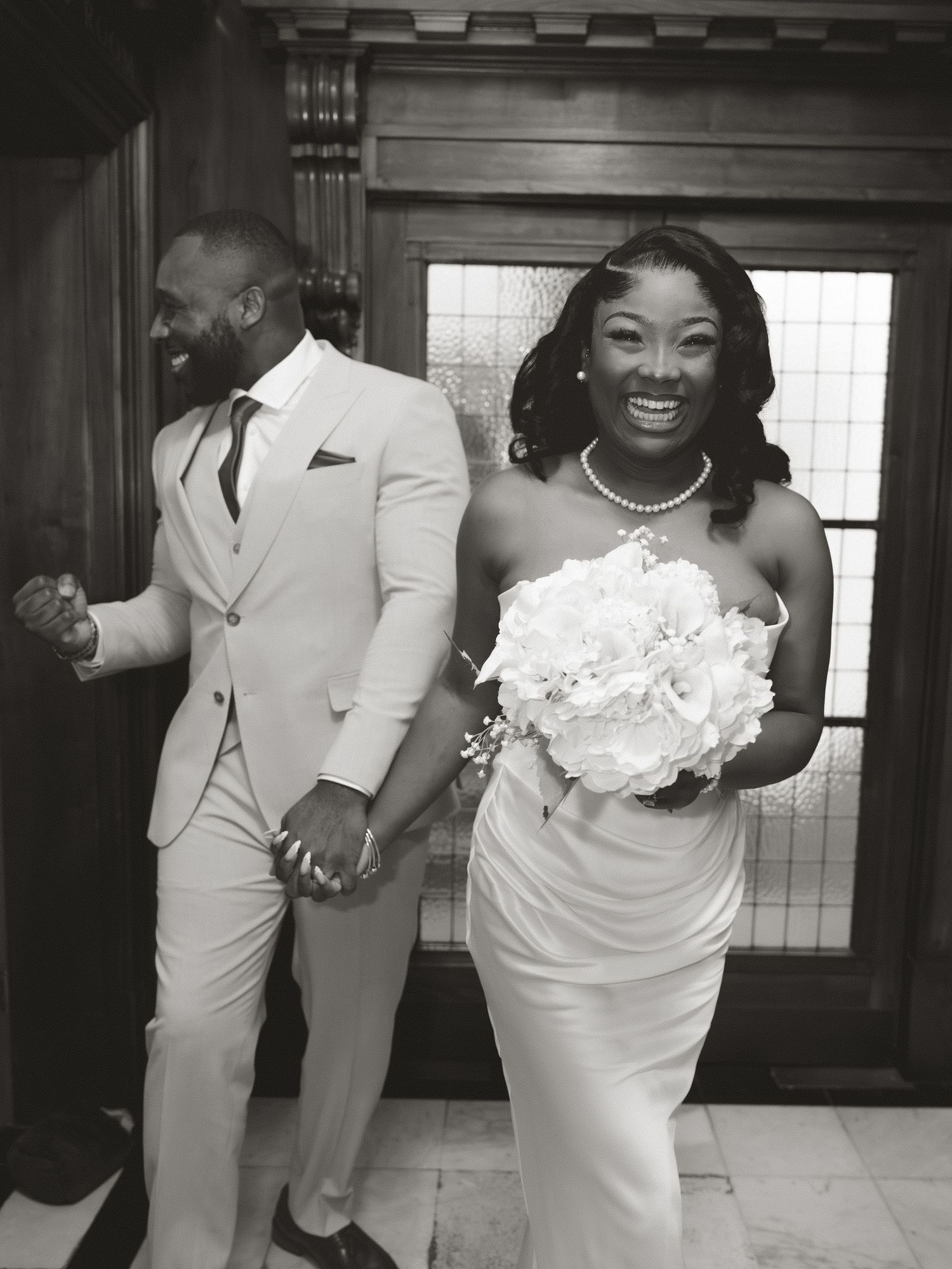 Black and white photo of a joyful bride in a white gown holding a bouquet and groom in a light suit holding hands.