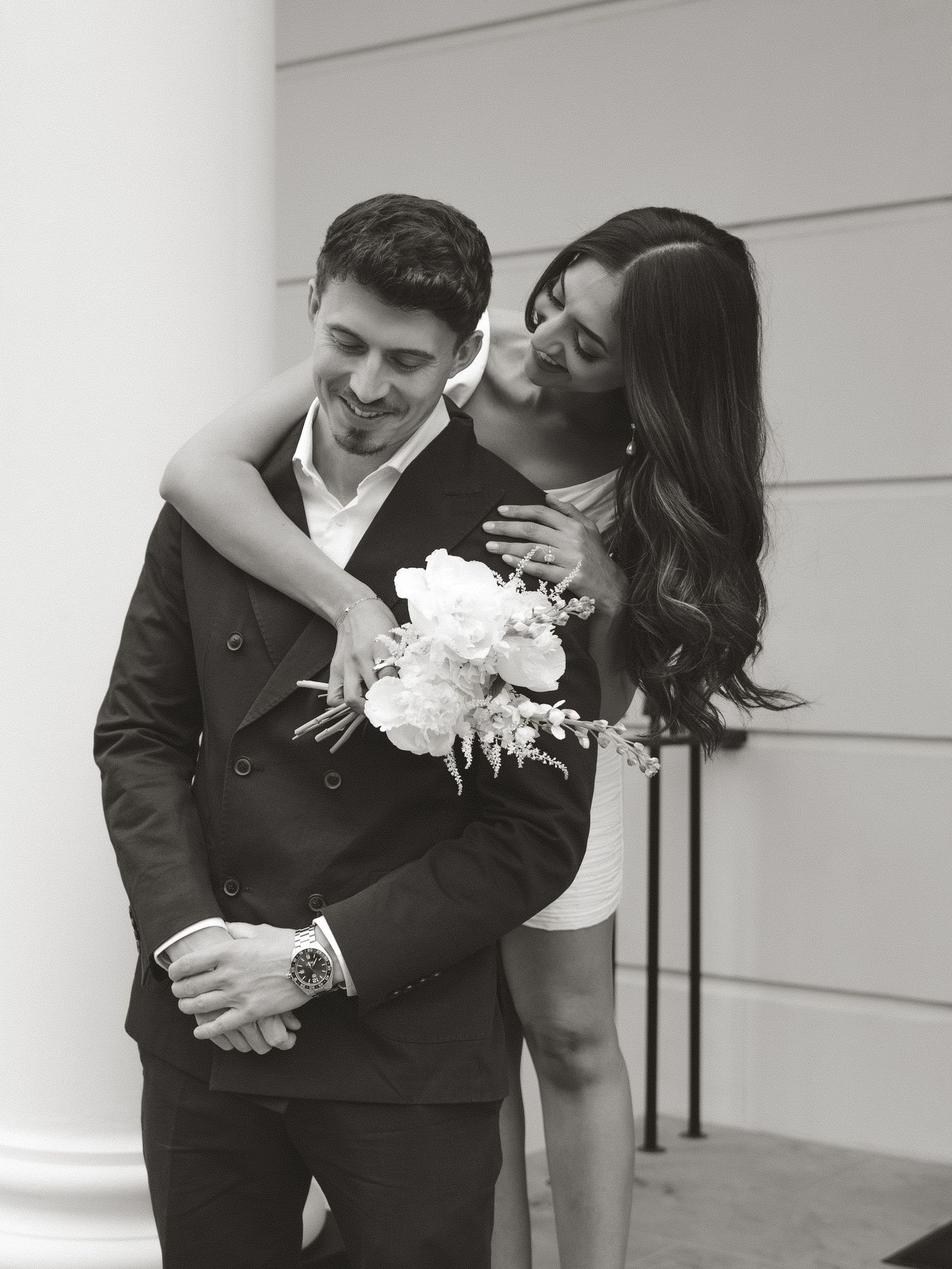 Black and white photo of a couple embracing, woman holding bouquet of flowers, man in suit smiling, romantic engagement portrait.