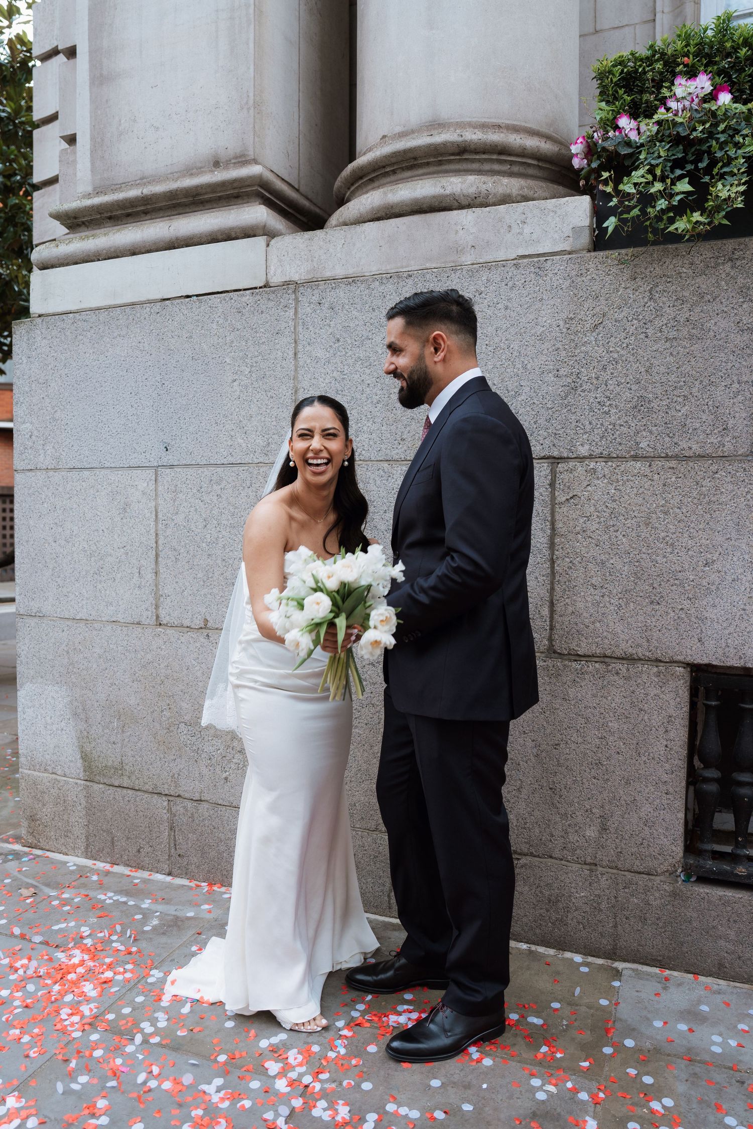 A bride and groom share a joyful moment together on their wedding day against a stone building wall.