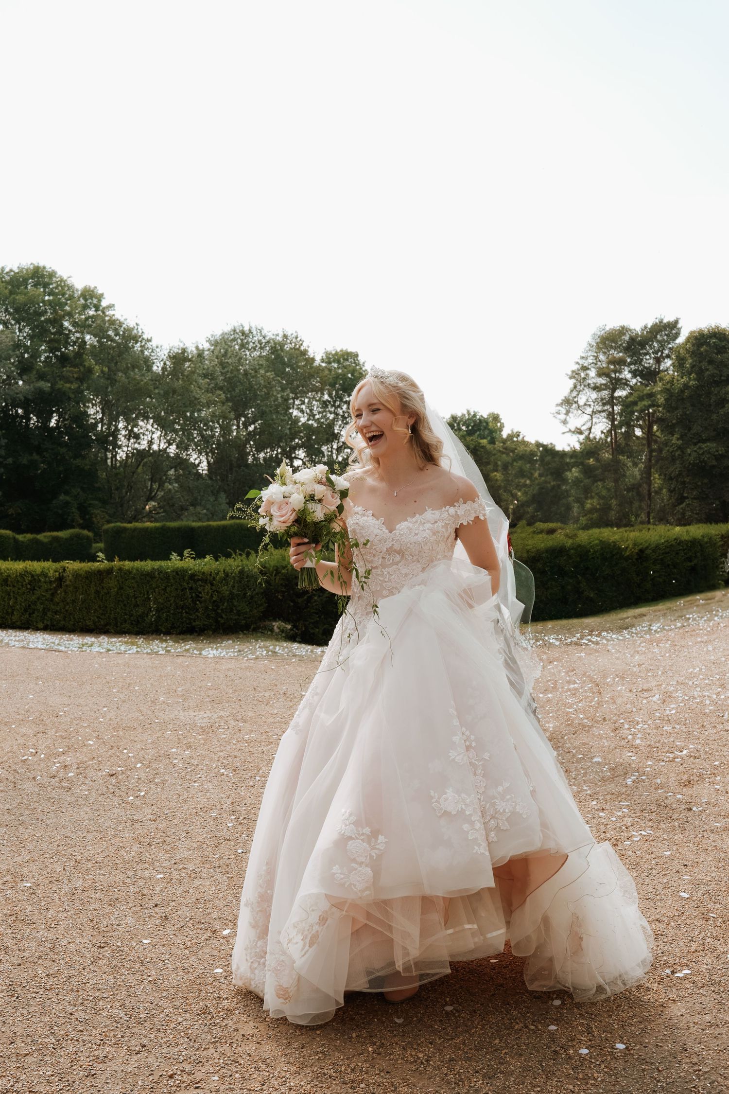 A flowing white wedding dress billows gracefully across a gravel pathway in an outdoor garden setting.