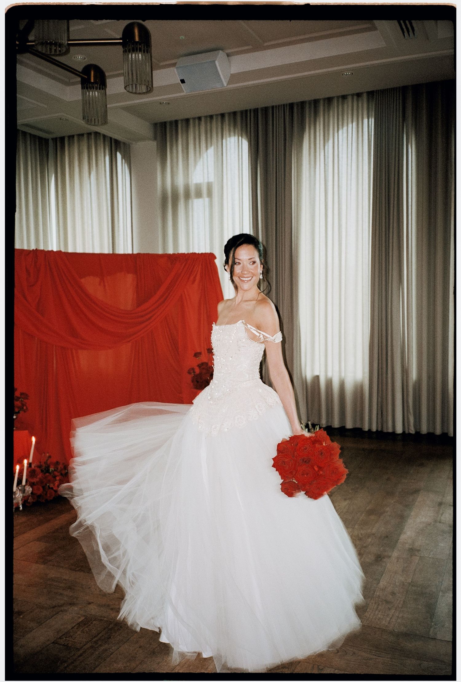 A bride in a flowing white ball gown poses against a red backdrop with matching red bouquet in an elegant venue.
