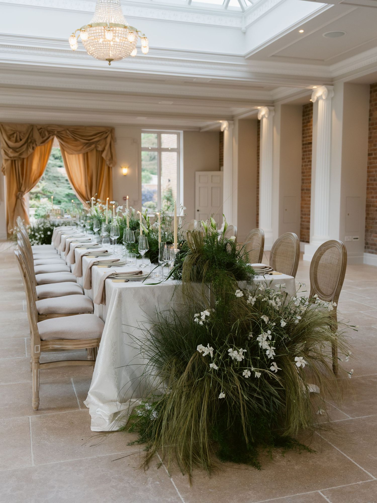 Elegant wedding reception table with lush greenery runner, white flowers, candles, gold chairs, and chandelier in a bright venue.