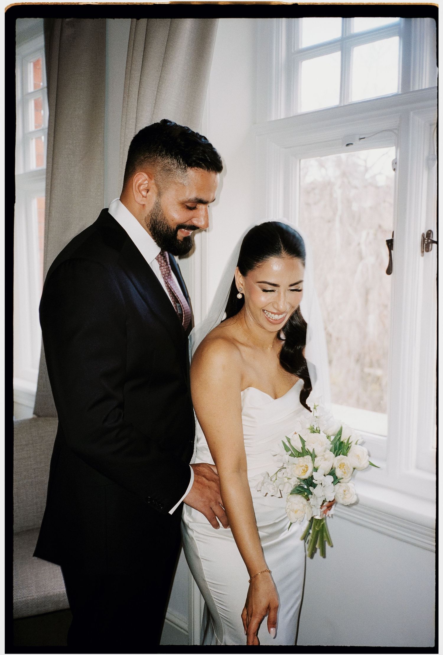 A romantic wedding couple shares intimate moments in a bright sunlit room with white walls and windows.