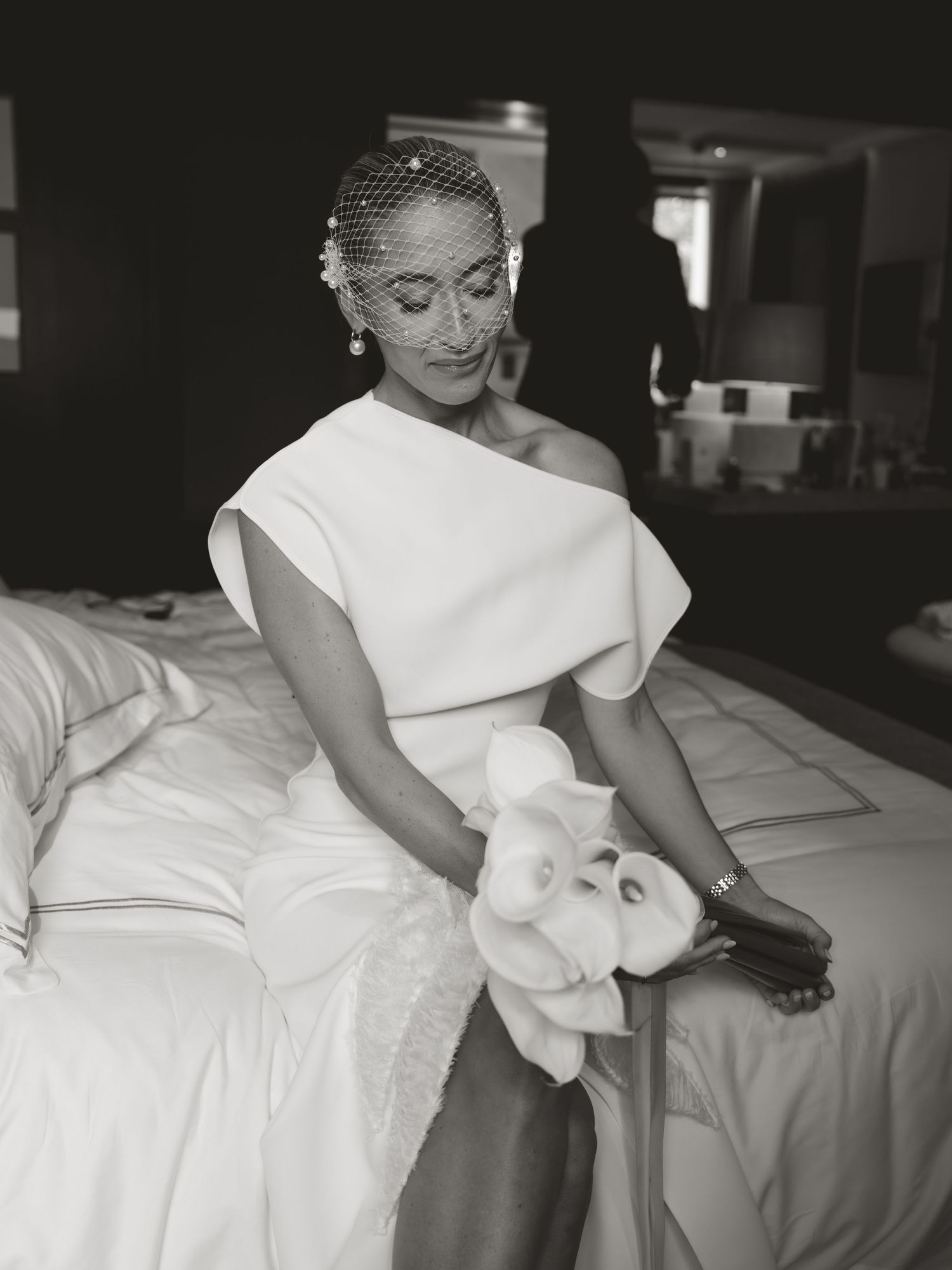 Elegant bride in one-shoulder white gown with birdcage veil holds floral bouquet while seated on bed in black and white photo.