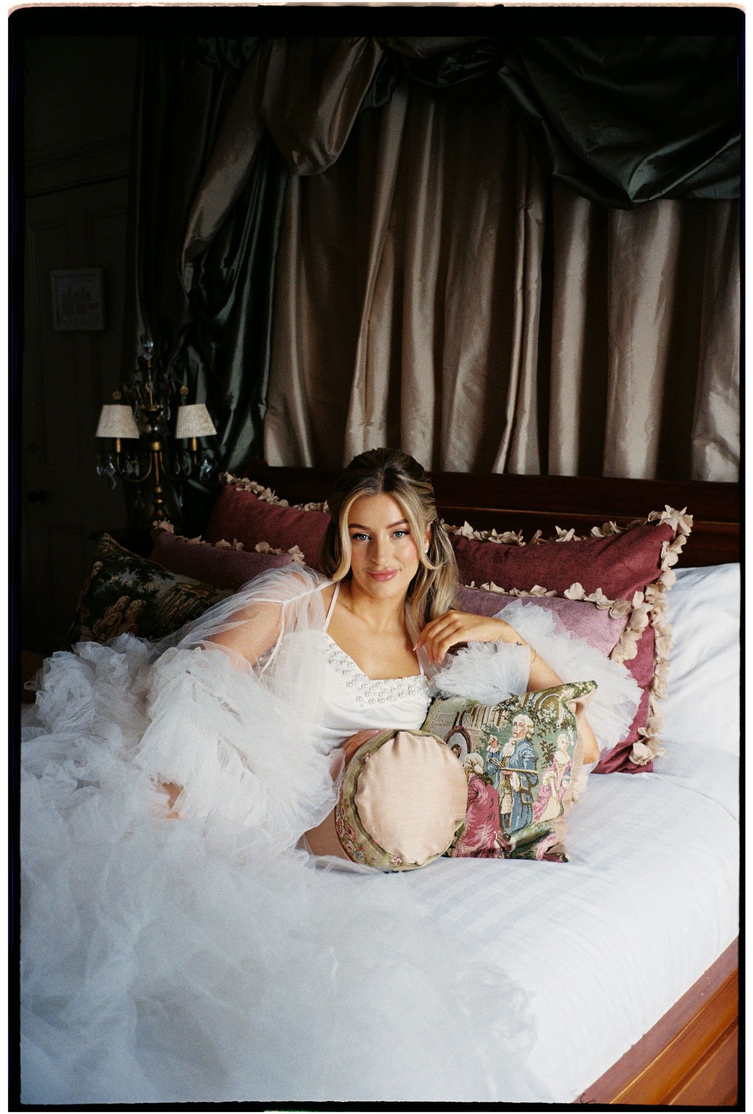 Bride in white tulle gown lounging on a luxurious bed with velvet pillows in an elegant dark bedroom.