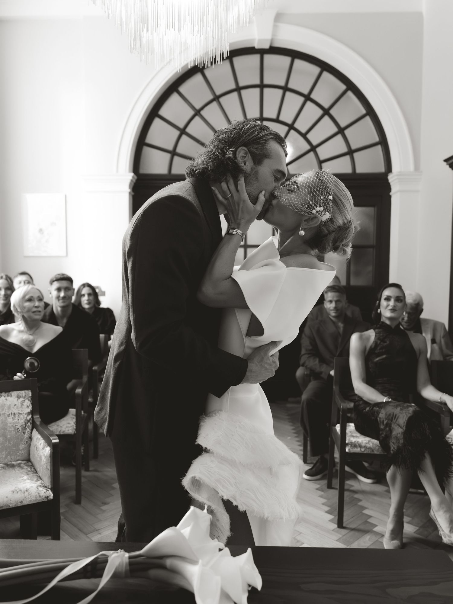 Black and white photo of a couple sharing their first kiss at a wedding ceremony with guests seated in the background.