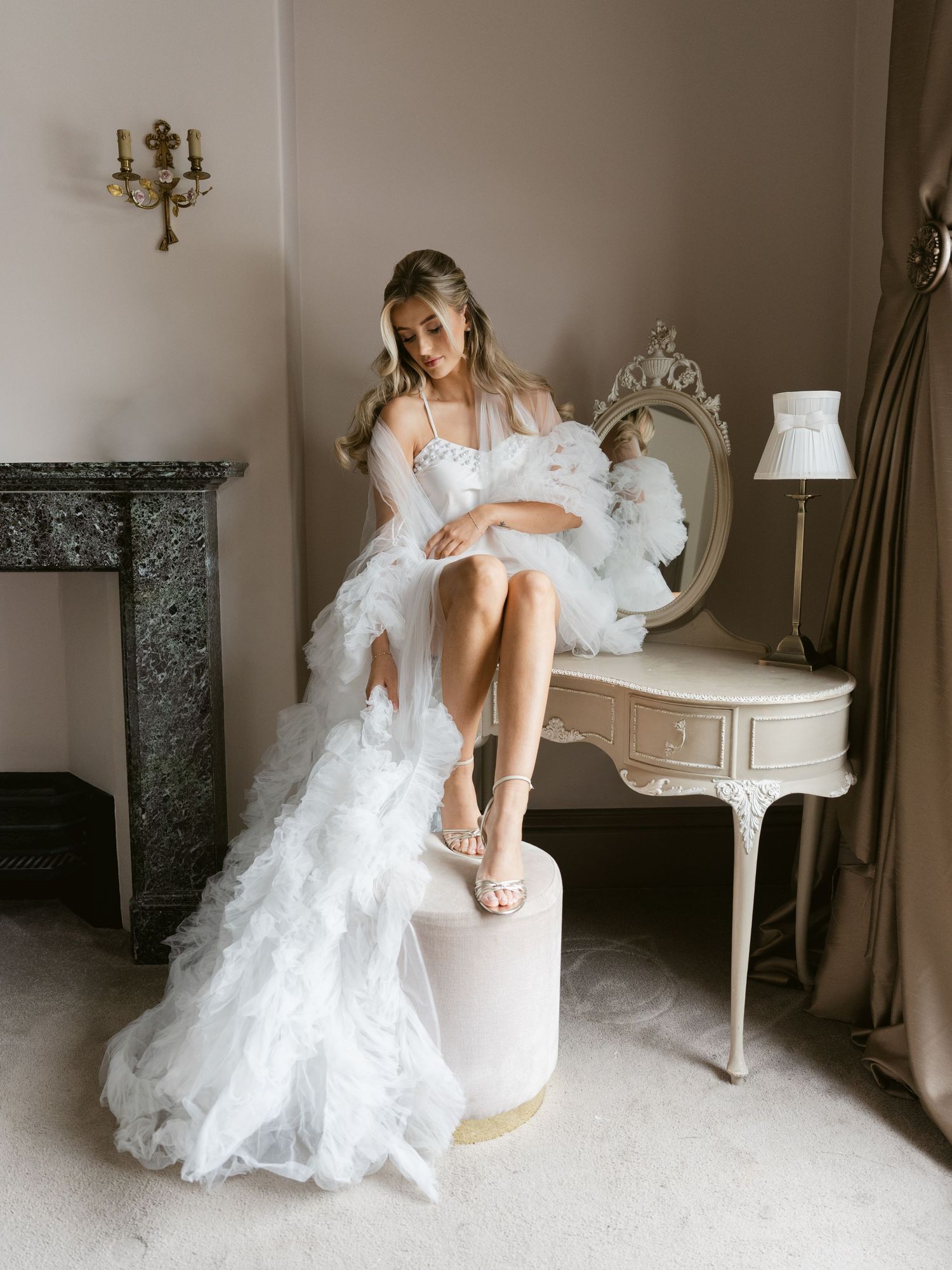Bride in a white ruffled tulle gown and sheer robe sits at a vintage vanity, adjusting her heels in an elegant room.