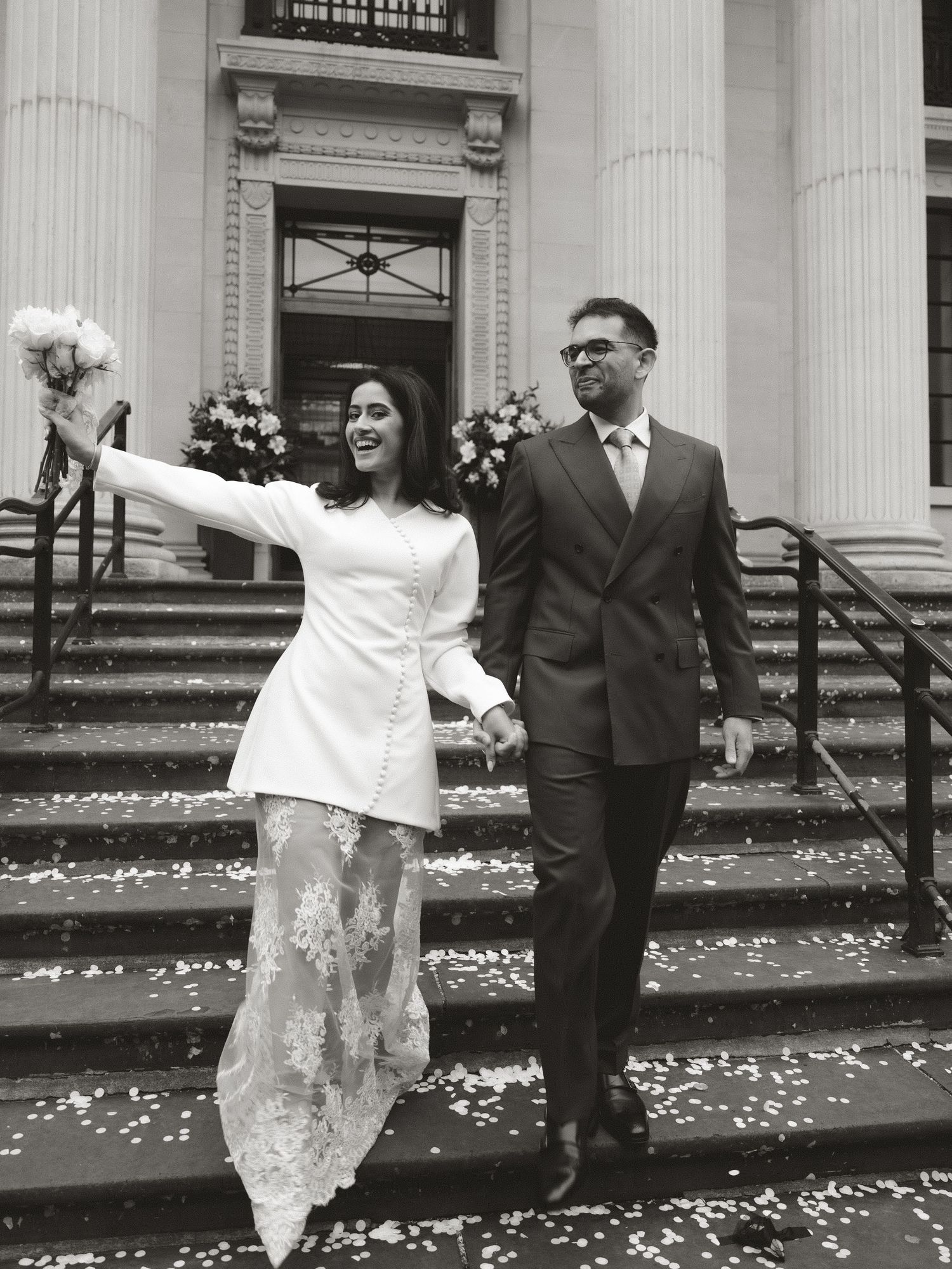 Wedding couple poses on steps of classical building with white columns and scattered flower petals.
