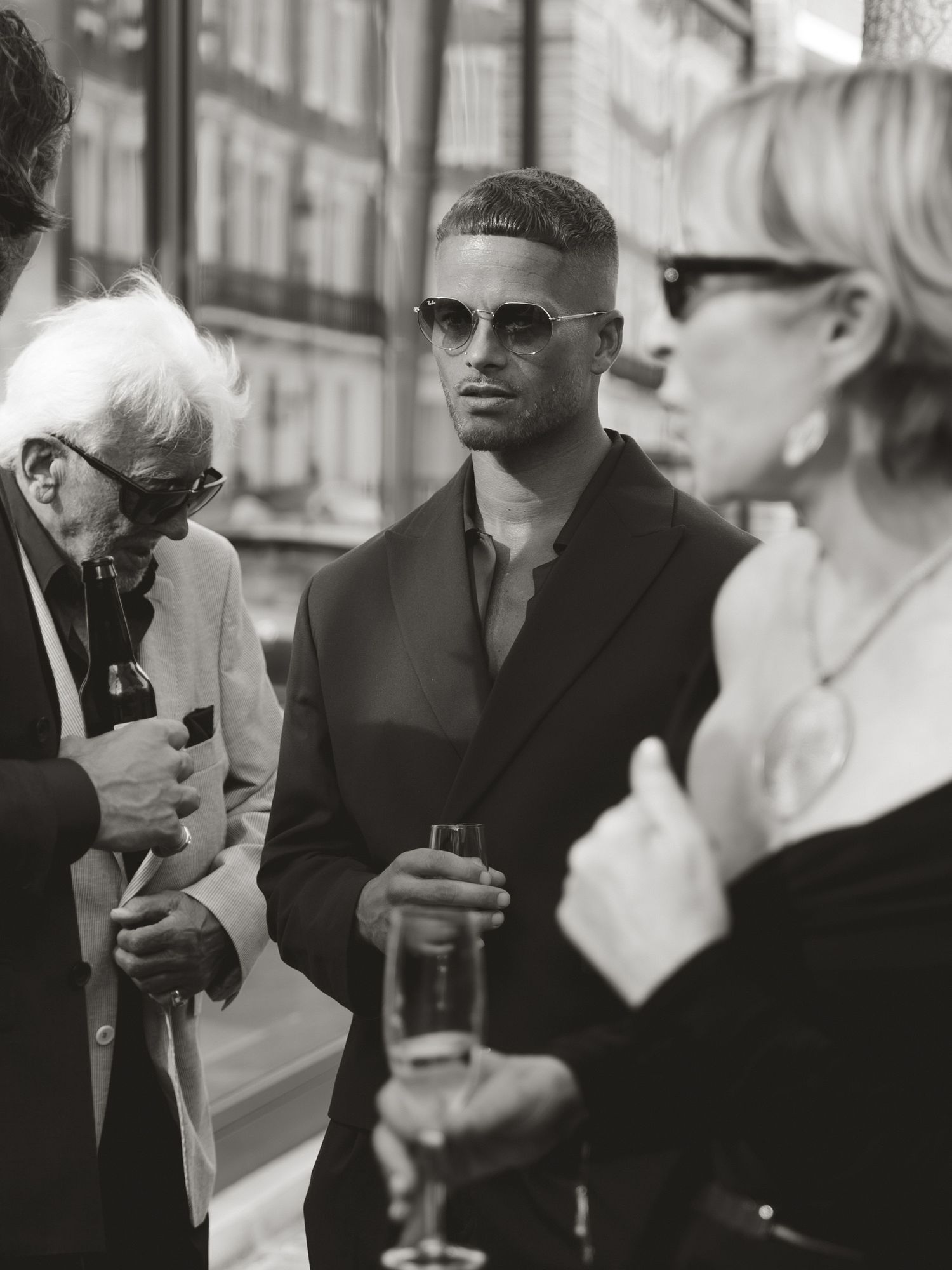 Black and white photo of stylish man in sunglasses and blazer holding drink at outdoor social gathering.
