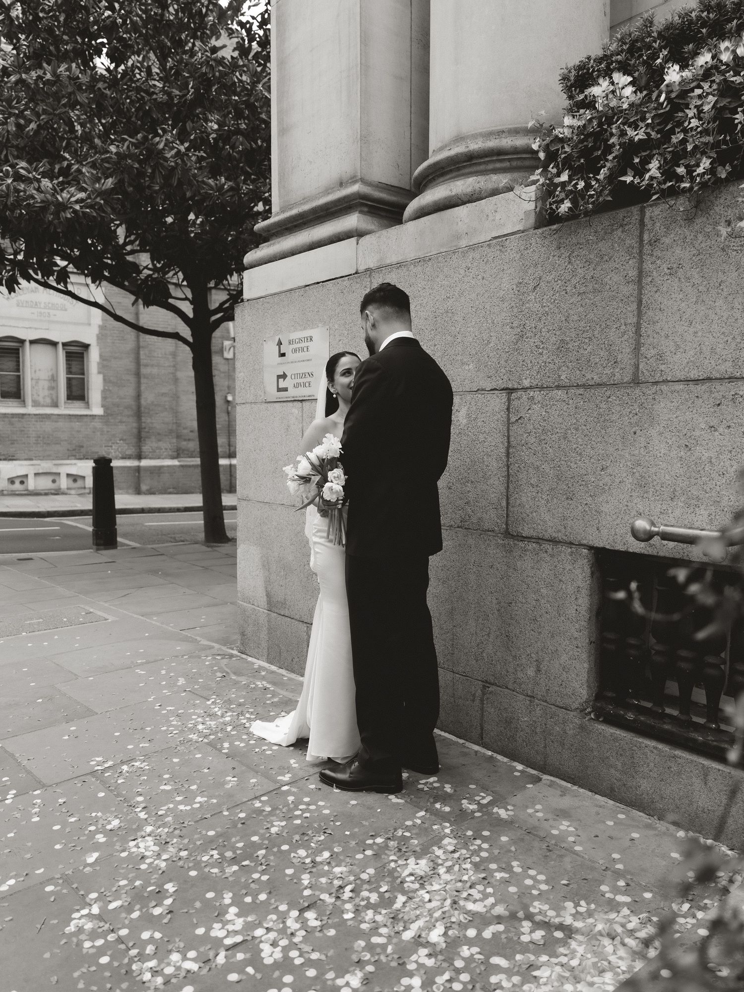 A black and white vintage wedding photograph showing a couple standing against a stone building with flower petals on the ground.