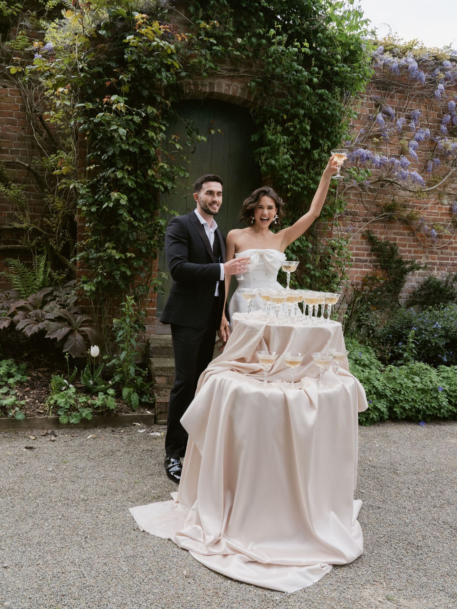 Bride and groom celebrate at champagne tower in lush garden wedding venue with ivy-covered brick wall backdrop.