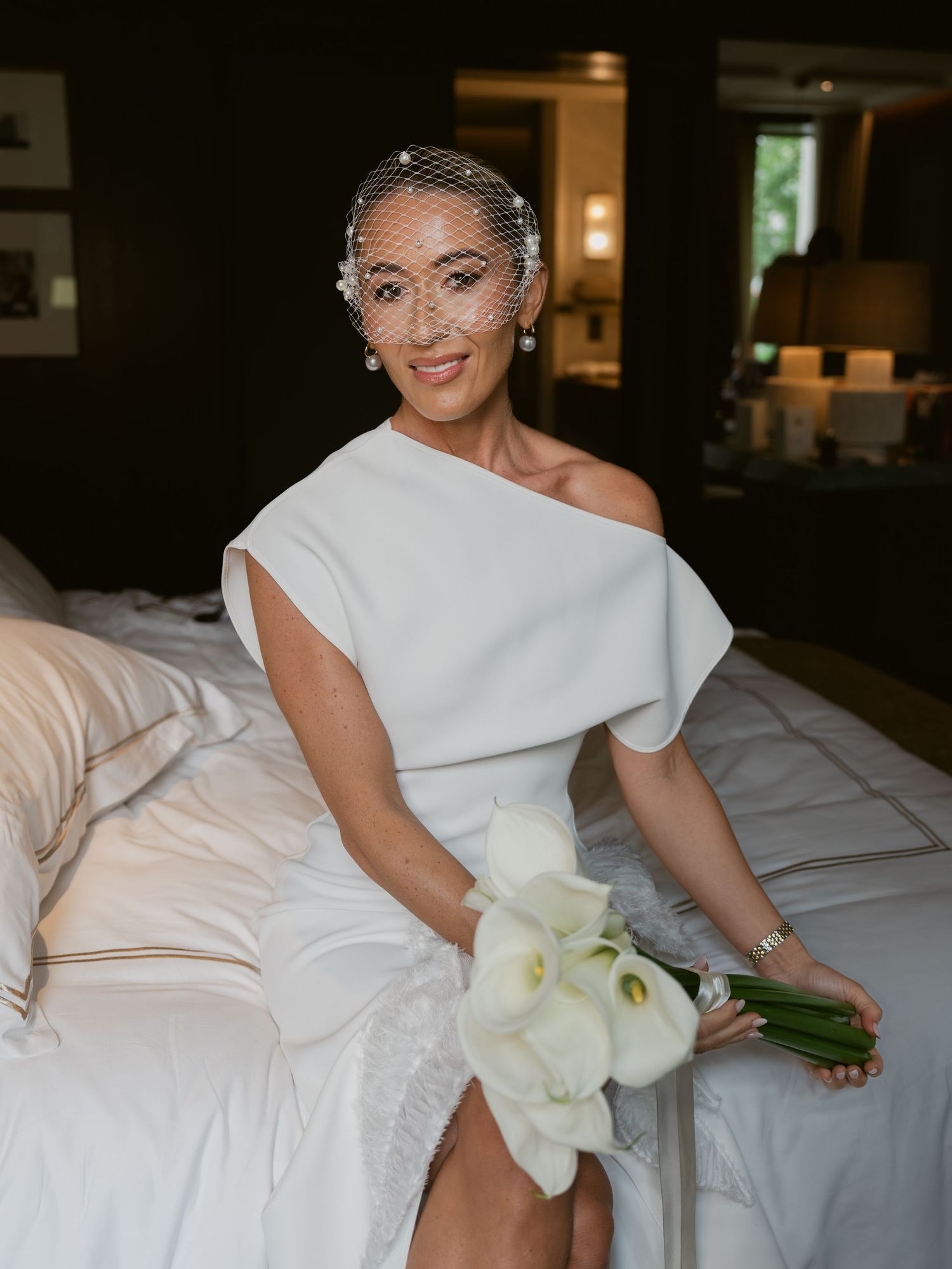 Elegant bride in white one-shoulder dress with birdcage veil holds white calla lily bouquet while sitting on bed.