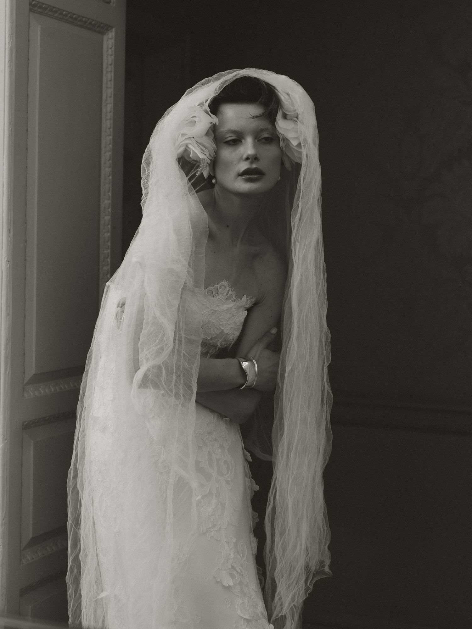 Black and white photo of a bride in lace wedding dress and long veil, posing elegantly in a doorway.