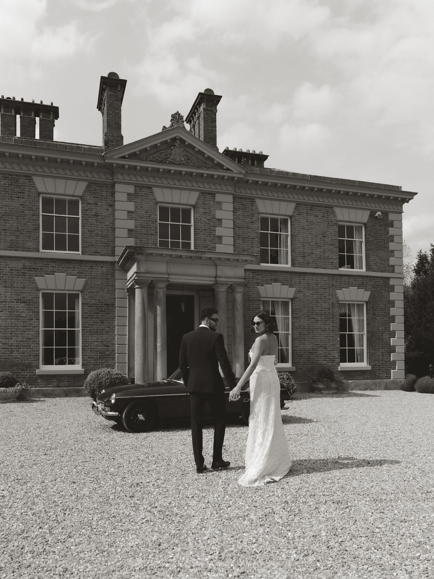 Black and white photo of a bride and groom holding hands before a grand Georgian brick manor house.