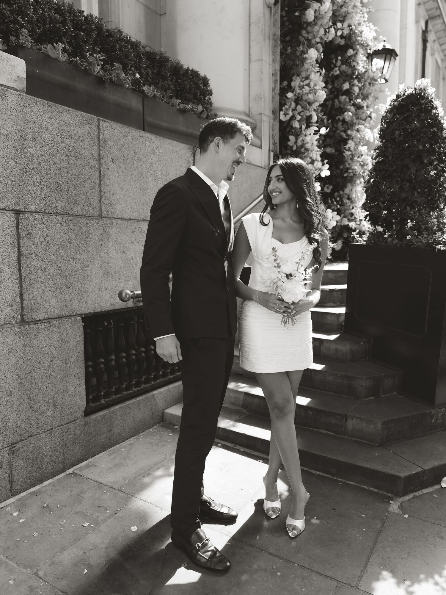 Black and white photo of a couple in wedding attire smiling at each other on a city sidewalk.