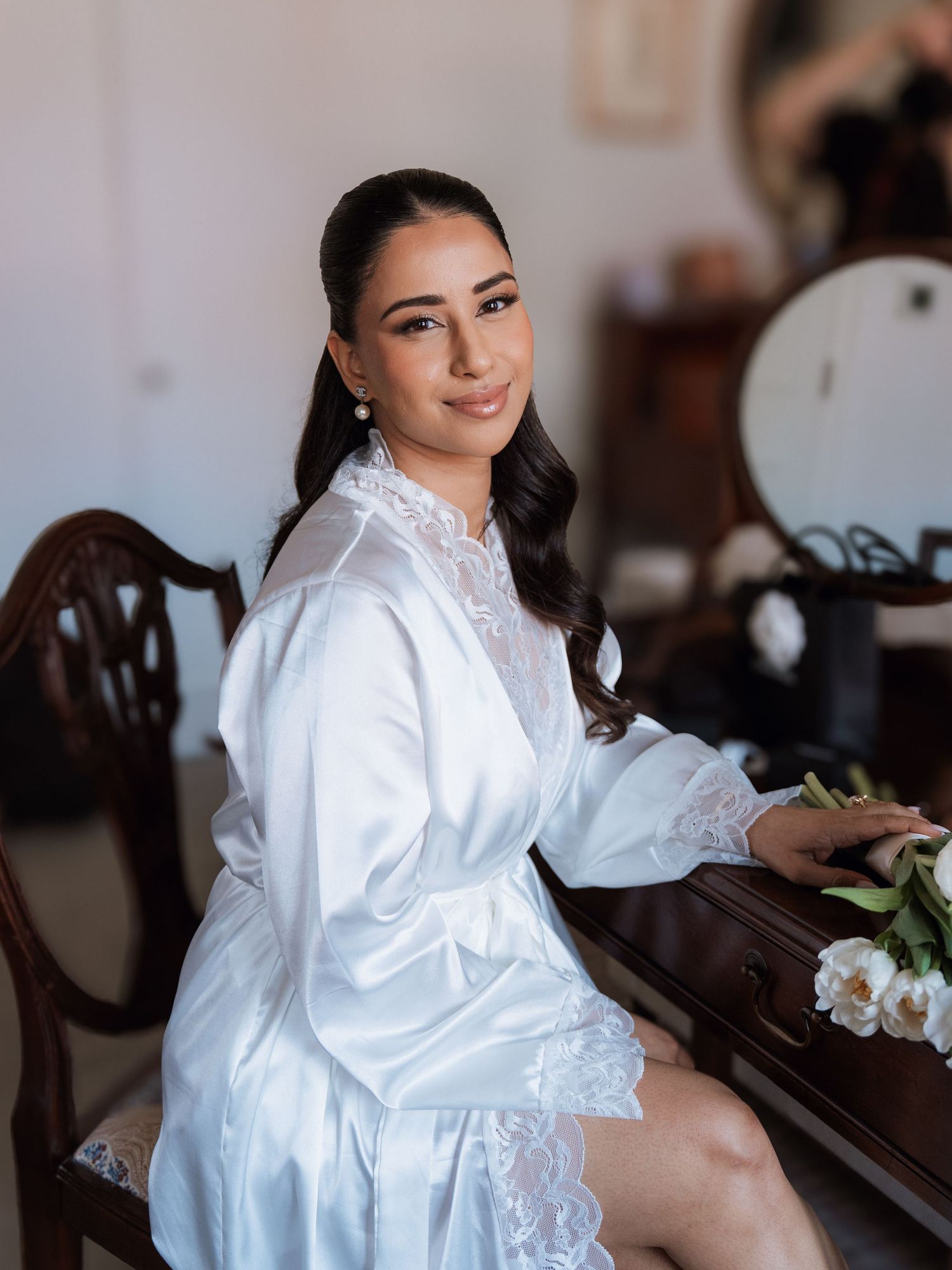 A person wearing a white satin robe sits elegantly in a traditional interior setting with wooden furniture.