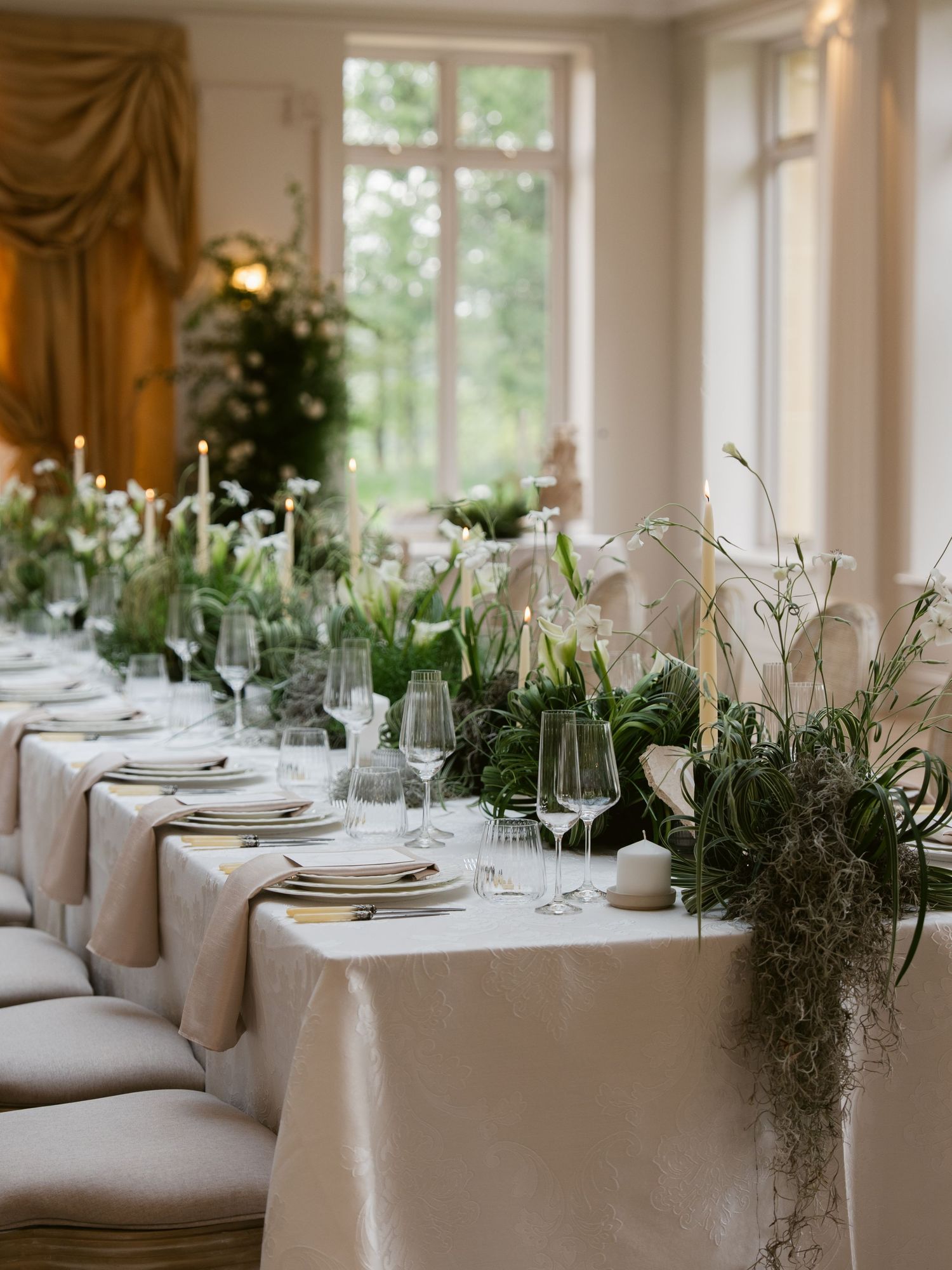 Elegant wedding reception table with white linen, greenery garland, white florals, and tall candles near sunlit windows.