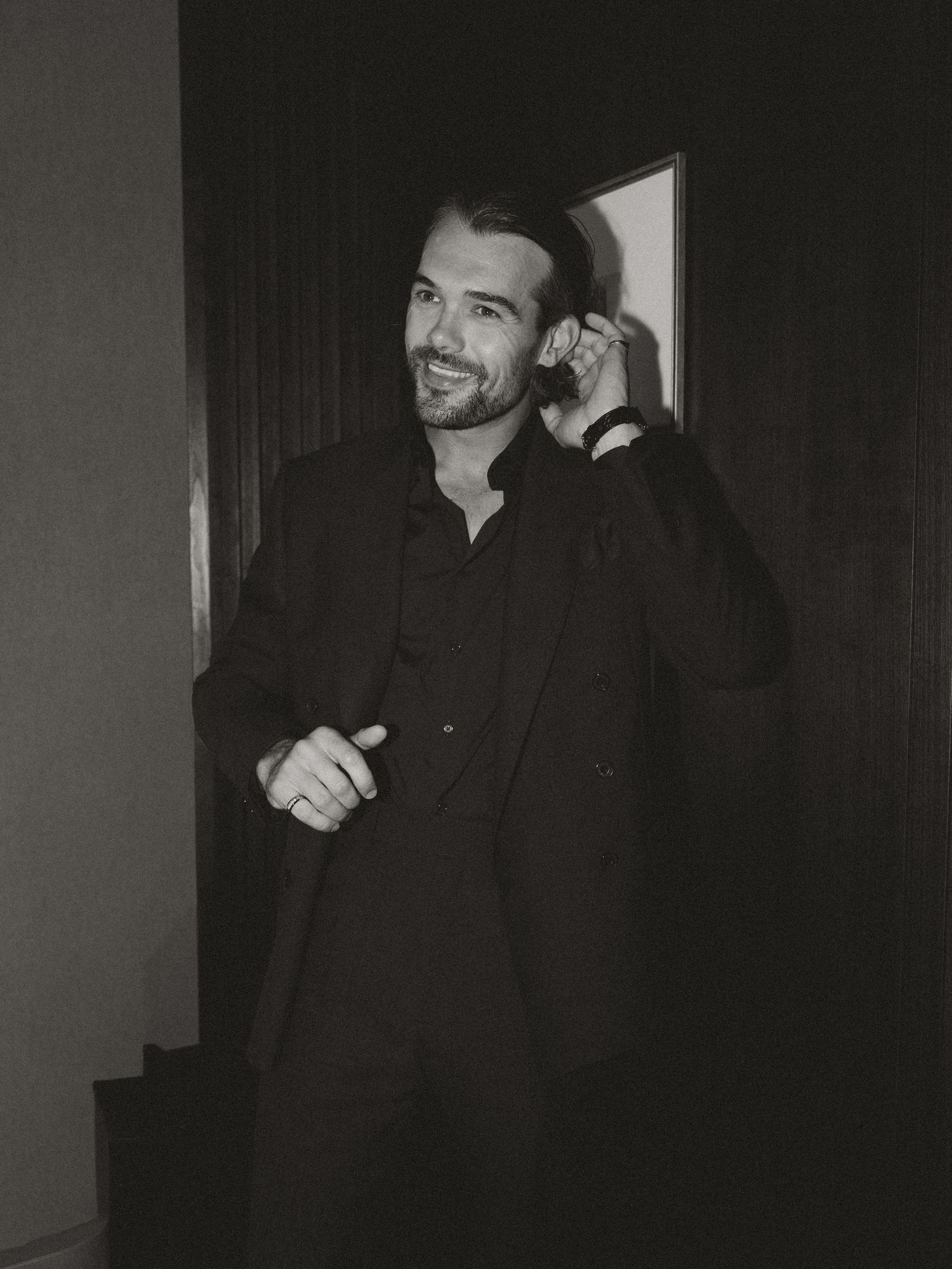 Black and white photo of a smiling man in a dark suit standing in a doorway, looking stylish and confident.