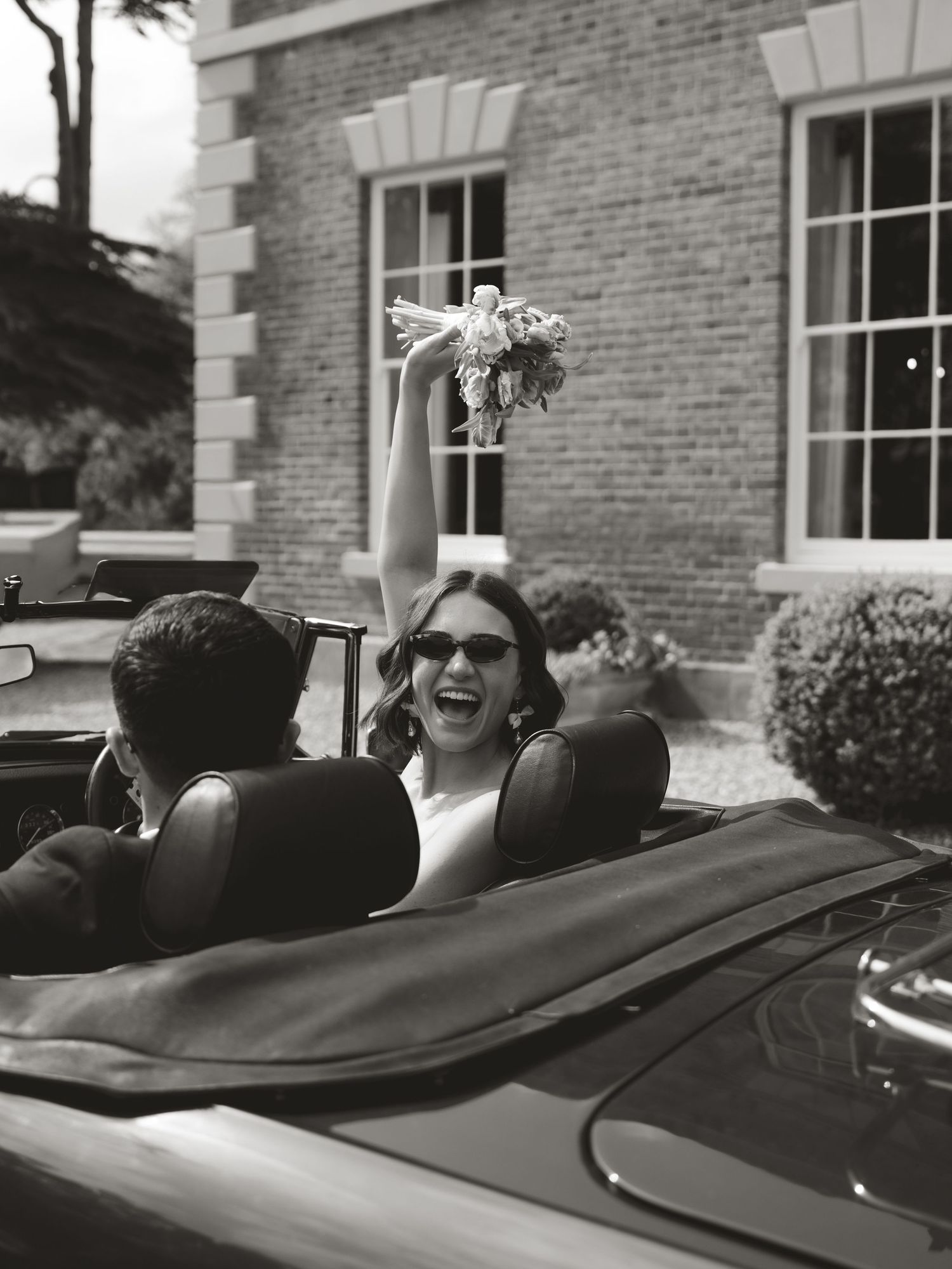 Black and white photo of a joyful bride raising her bouquet in a convertible car outside a brick manor house.