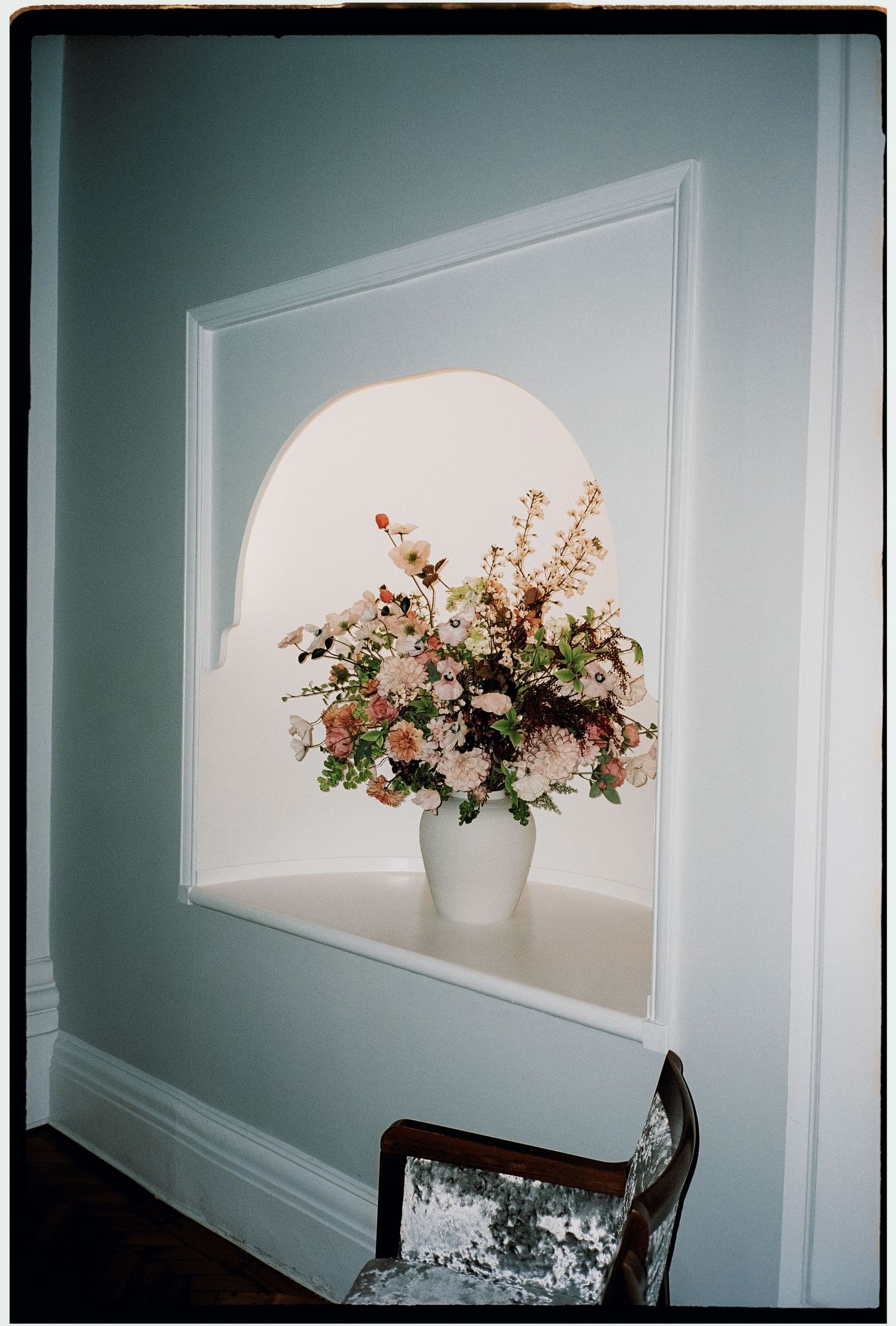 A delicate floral arrangement sits in a white vase within an arched wall niche against a sage green wall.