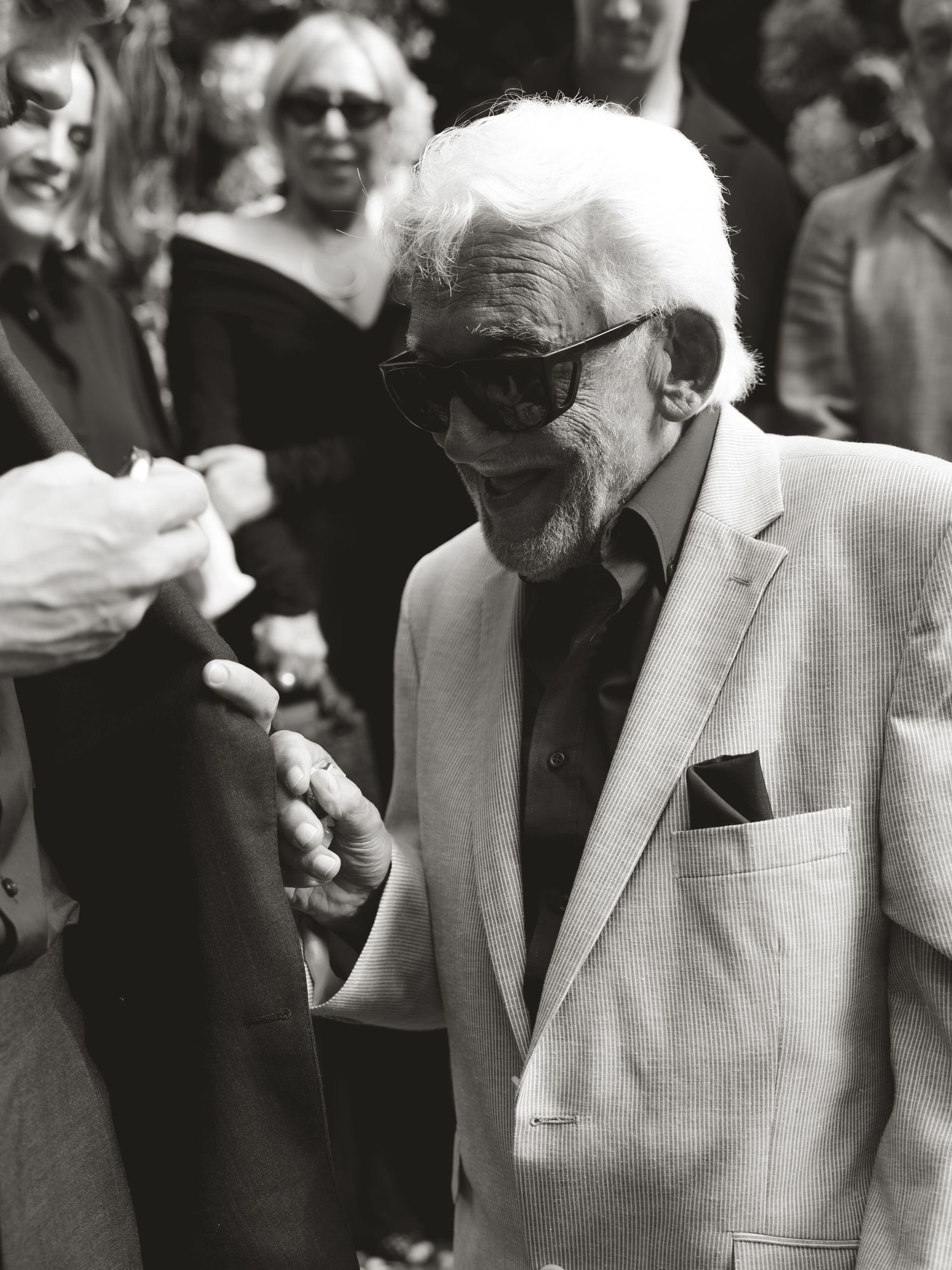 Elderly man in light suit and sunglasses shaking hands at outdoor event, black and white photo.