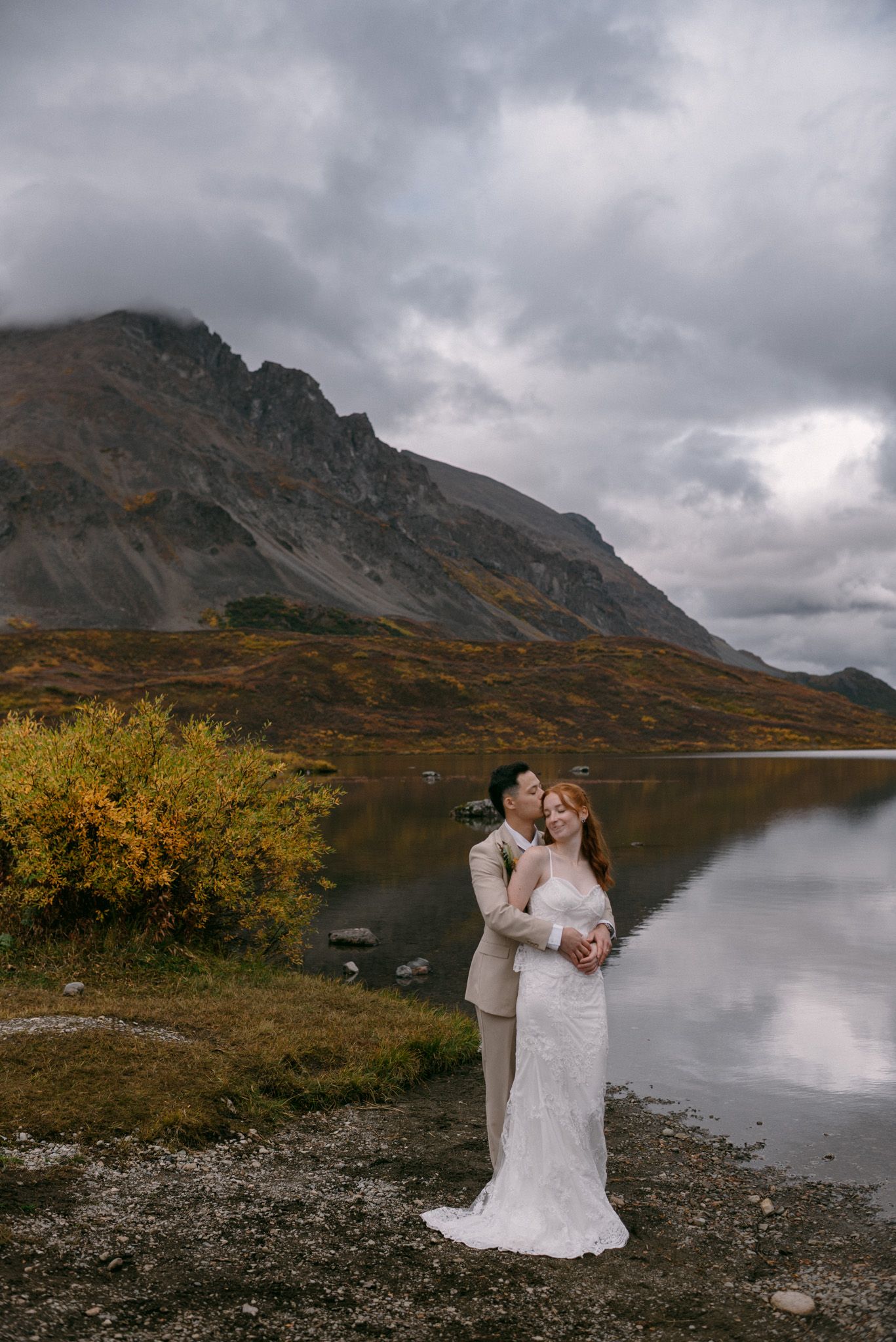 Bride and groom eloping on Denali Highway in Alaska, sharing an intimate moment surrounded by mountains and water.