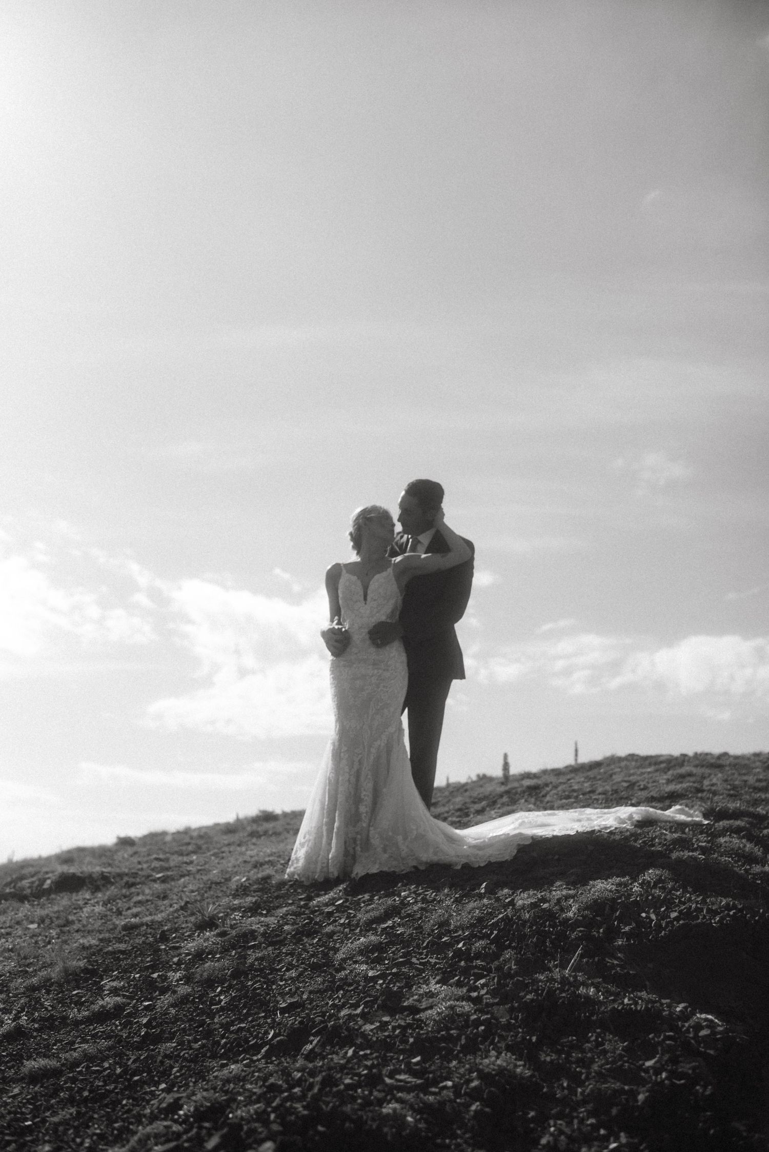 Black and white portrait of a couple on a mountain peak in Bozeman, Montana, embracing under a sunny sky.