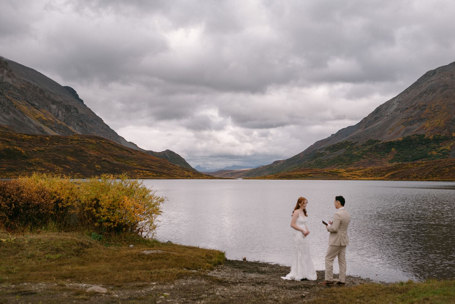 Couple exchanging wedding vows during an intimate Denali National Park elopement in Alaska, beside a calm lake with autumn mountain scenery.