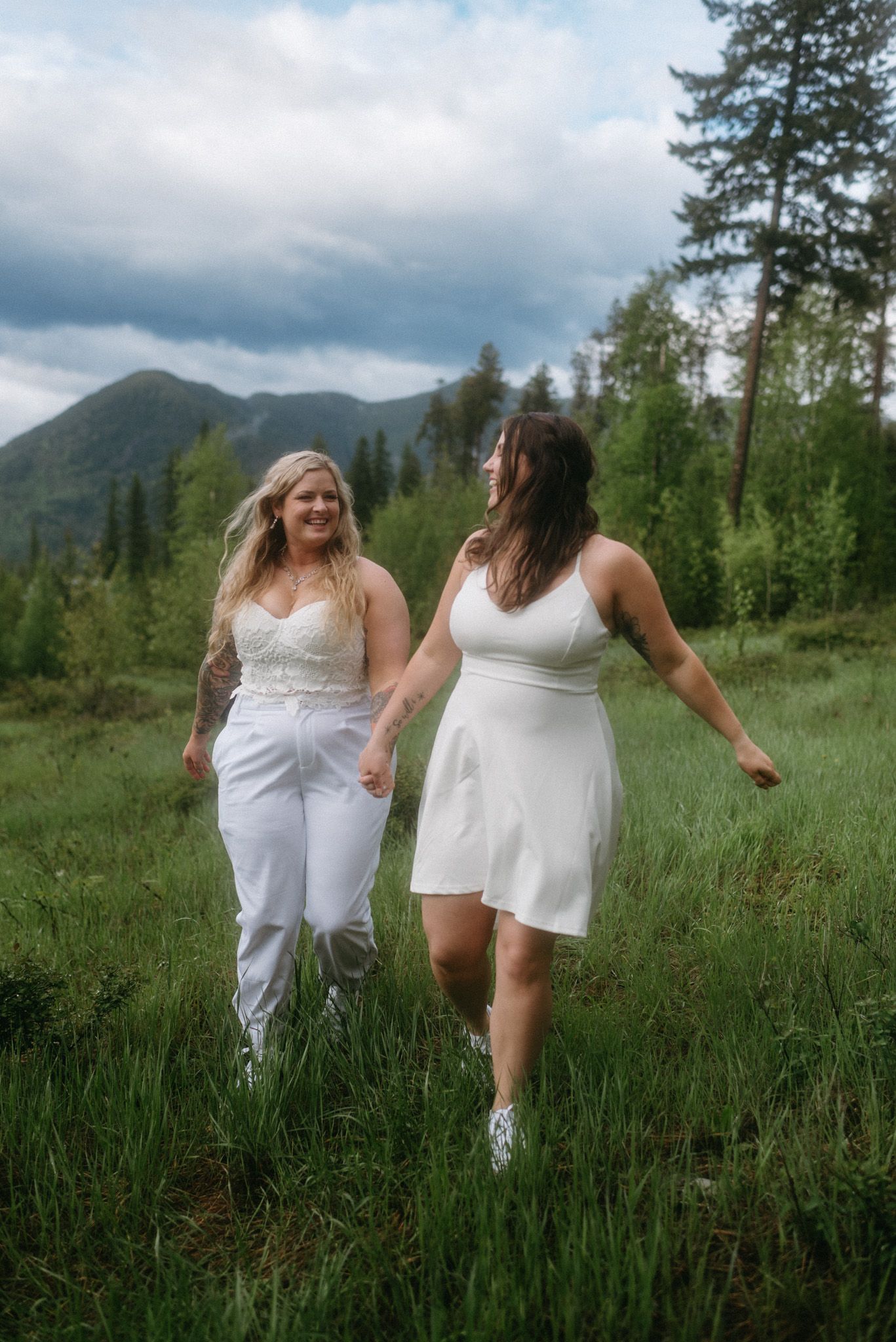 Joyful brides running together through green grass during their second wedding outfit shoot at Paddle Ridge, Glacier National Park.
