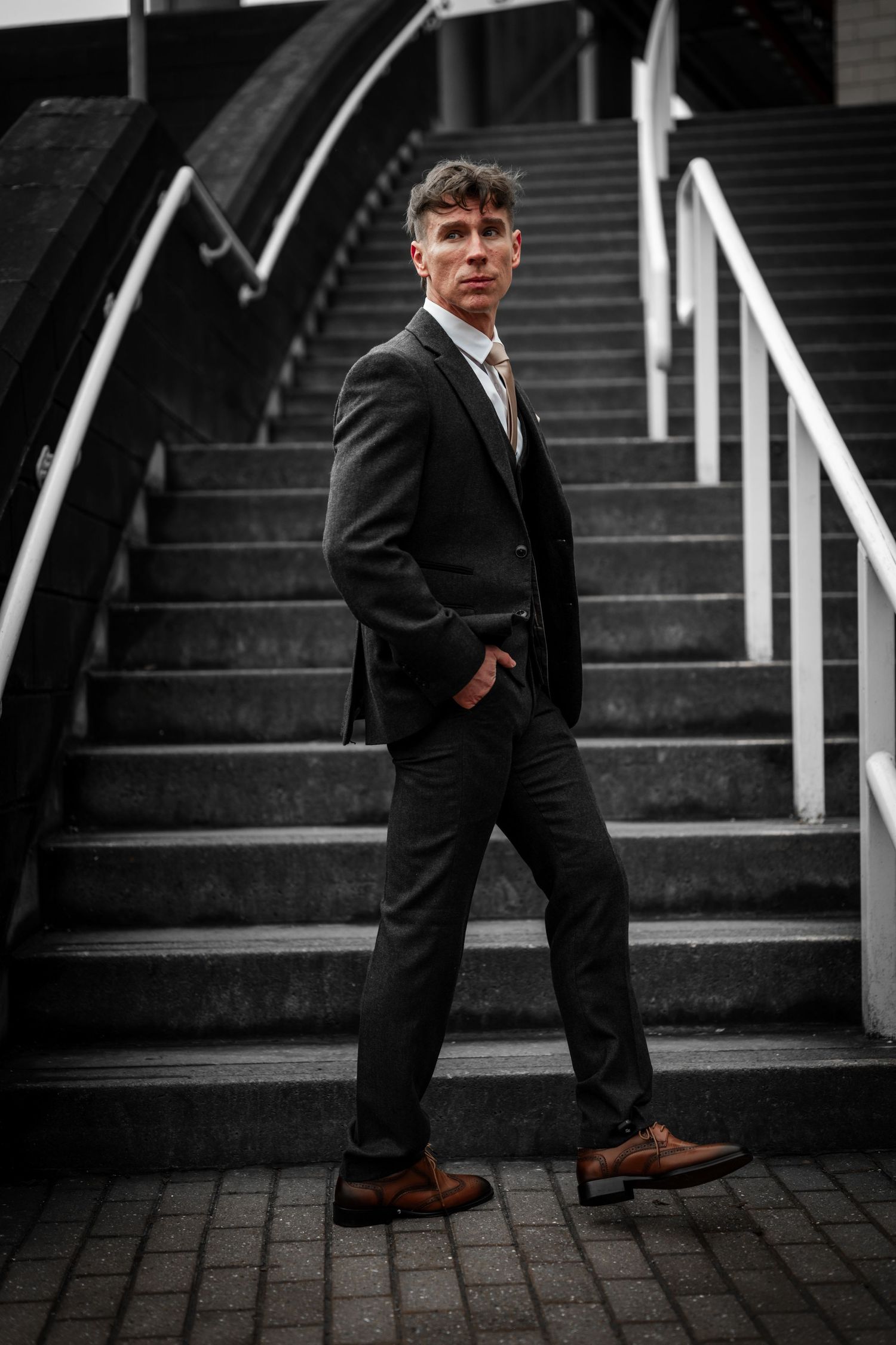 A person in a dark suit and brown shoes poses confidently on concrete steps with metal railings in moody lighting.
