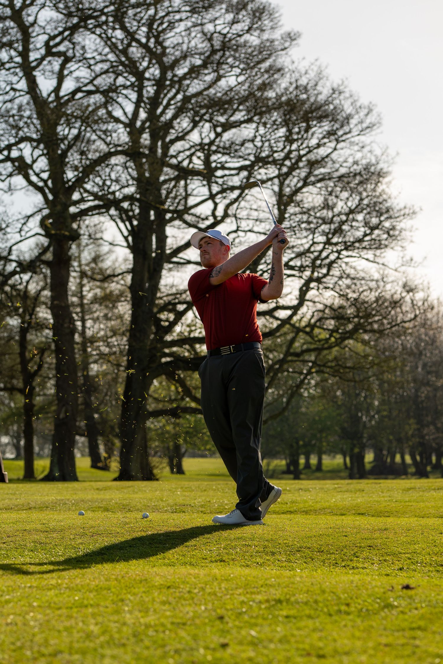 Golfer in red shirt and white cap completing a follow-through swing on a lush green course with bare trees in background.