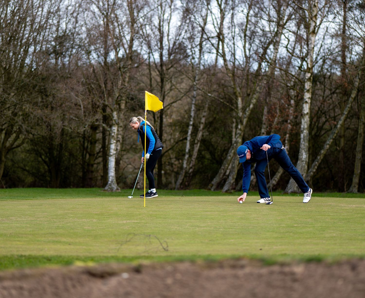 Two golfers on a lush green golf course near a yellow flag, with trees in the background on a cloudy day.