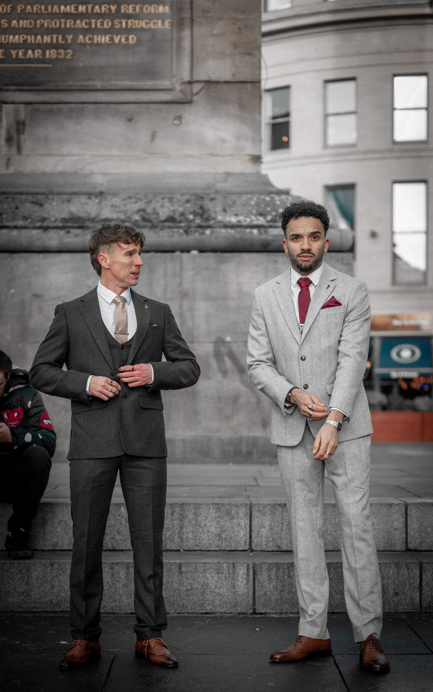 Two people in contrasting suits, one dark and one light, pose against an industrial concrete wall setting.