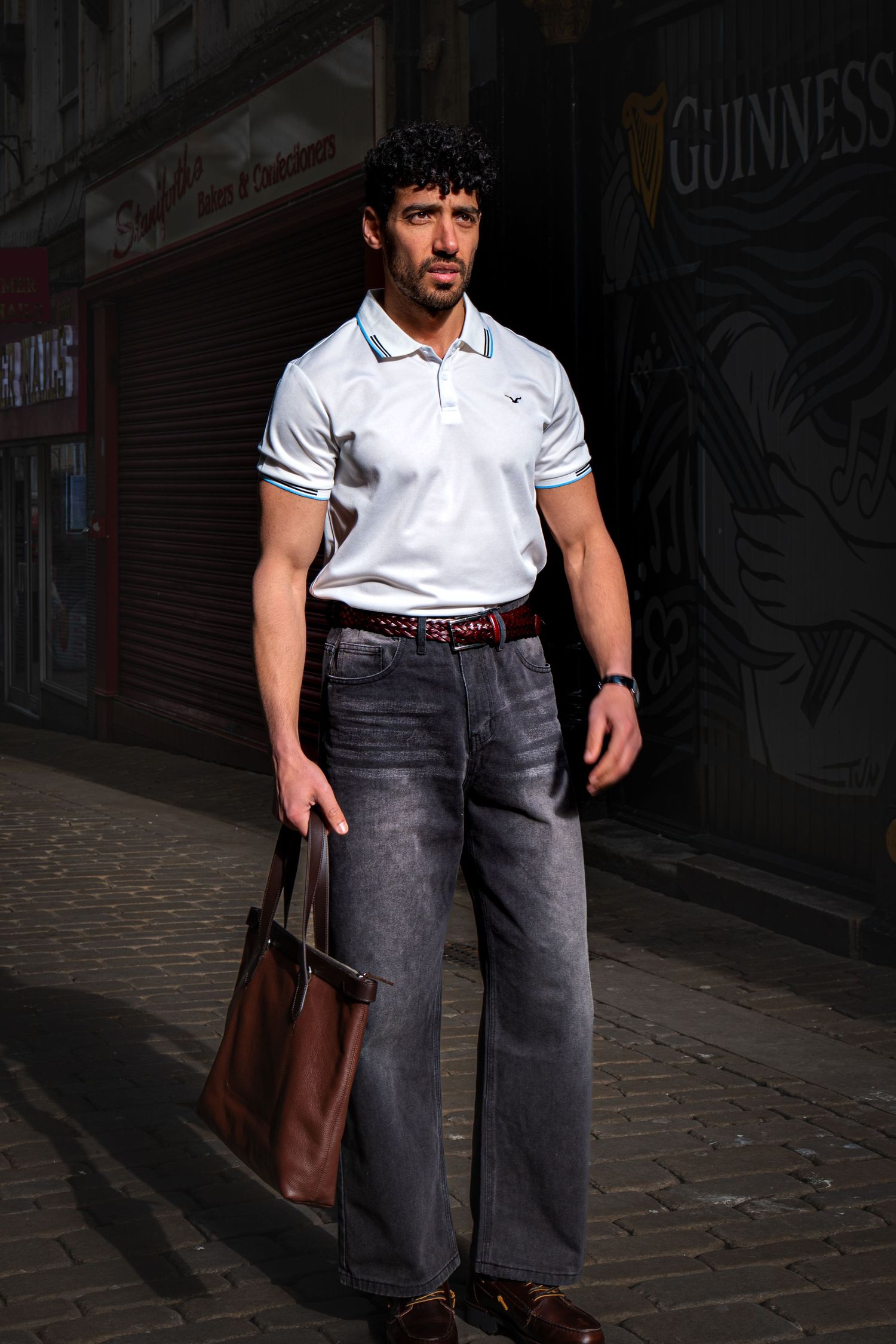 Man in white polo shirt and gray wide-leg jeans holding brown leather tote bag on cobblestone street.