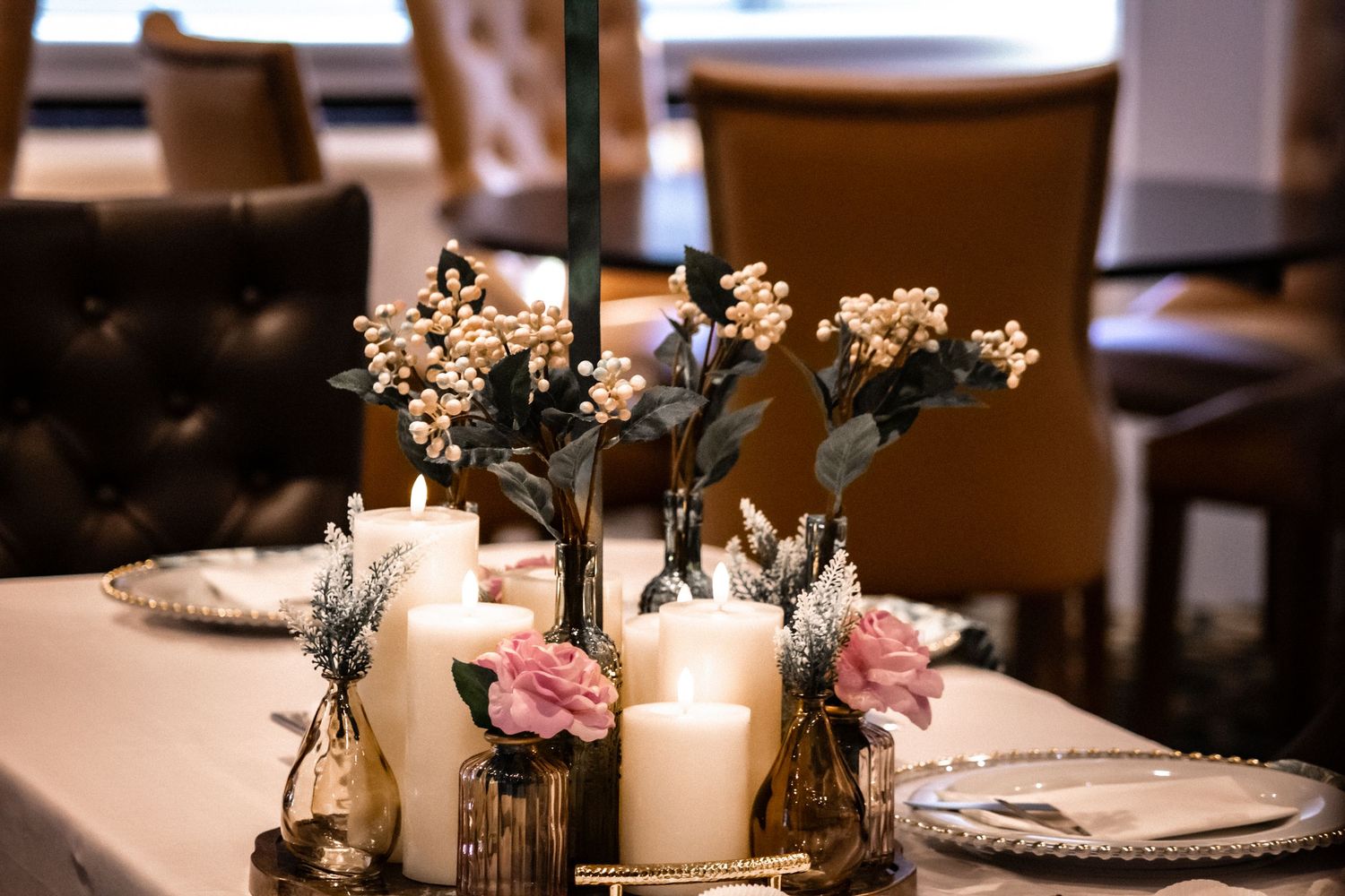 Elegant wedding reception table settings with white candles, pink roses, and delicate floral arrangements in glass vases.