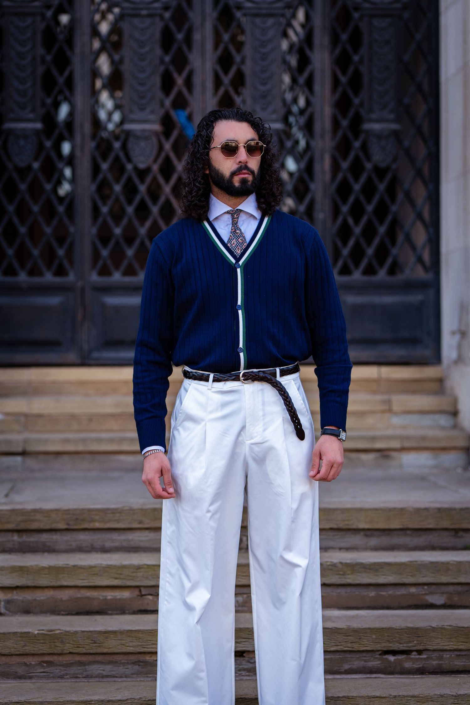 Stylish man in navy cardigan, white trousers, and round sunglasses standing on stone steps.